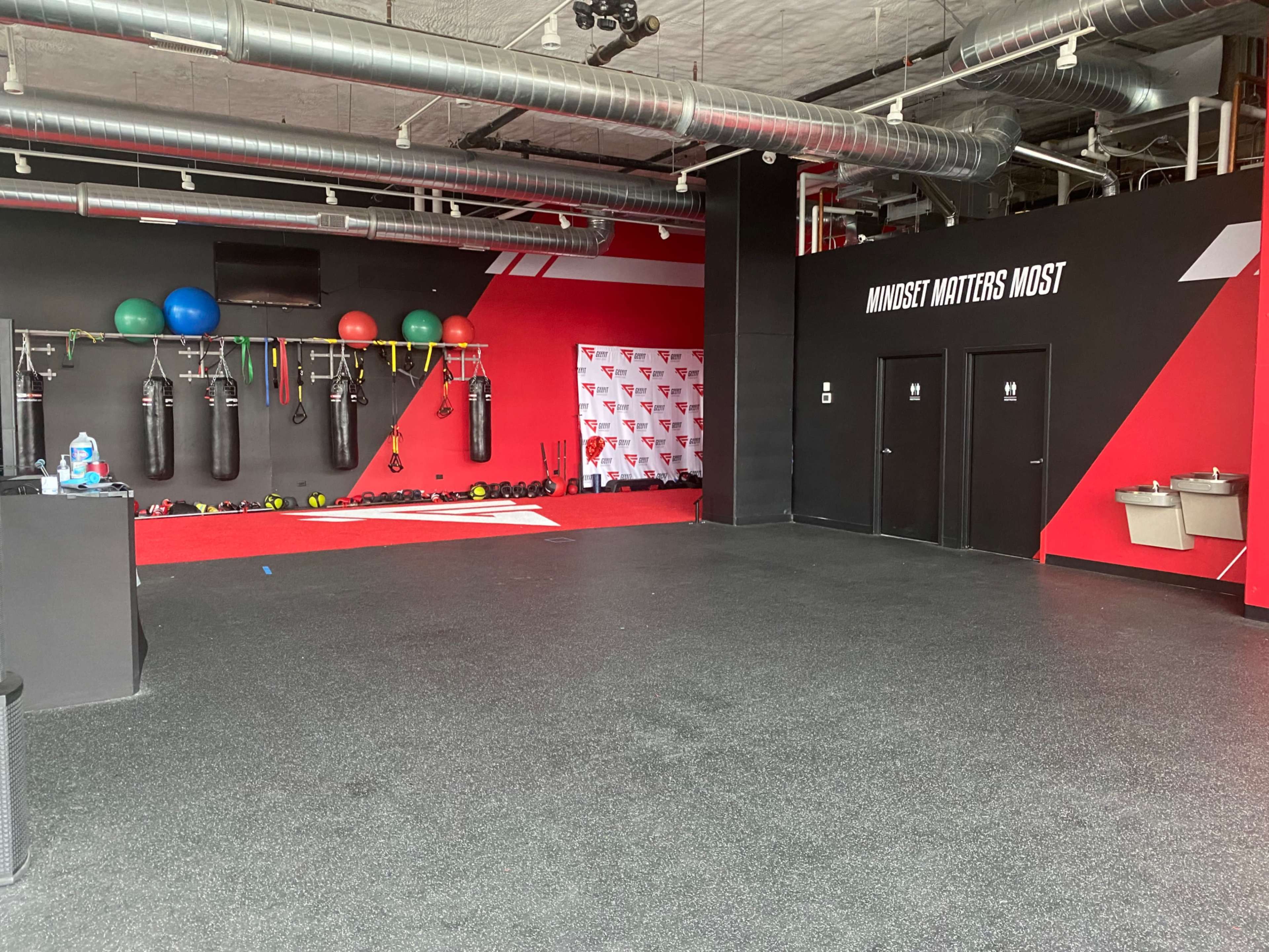 An empty gym with boxing bags, exercise balls, and weight equipment against a red and black backdrop.