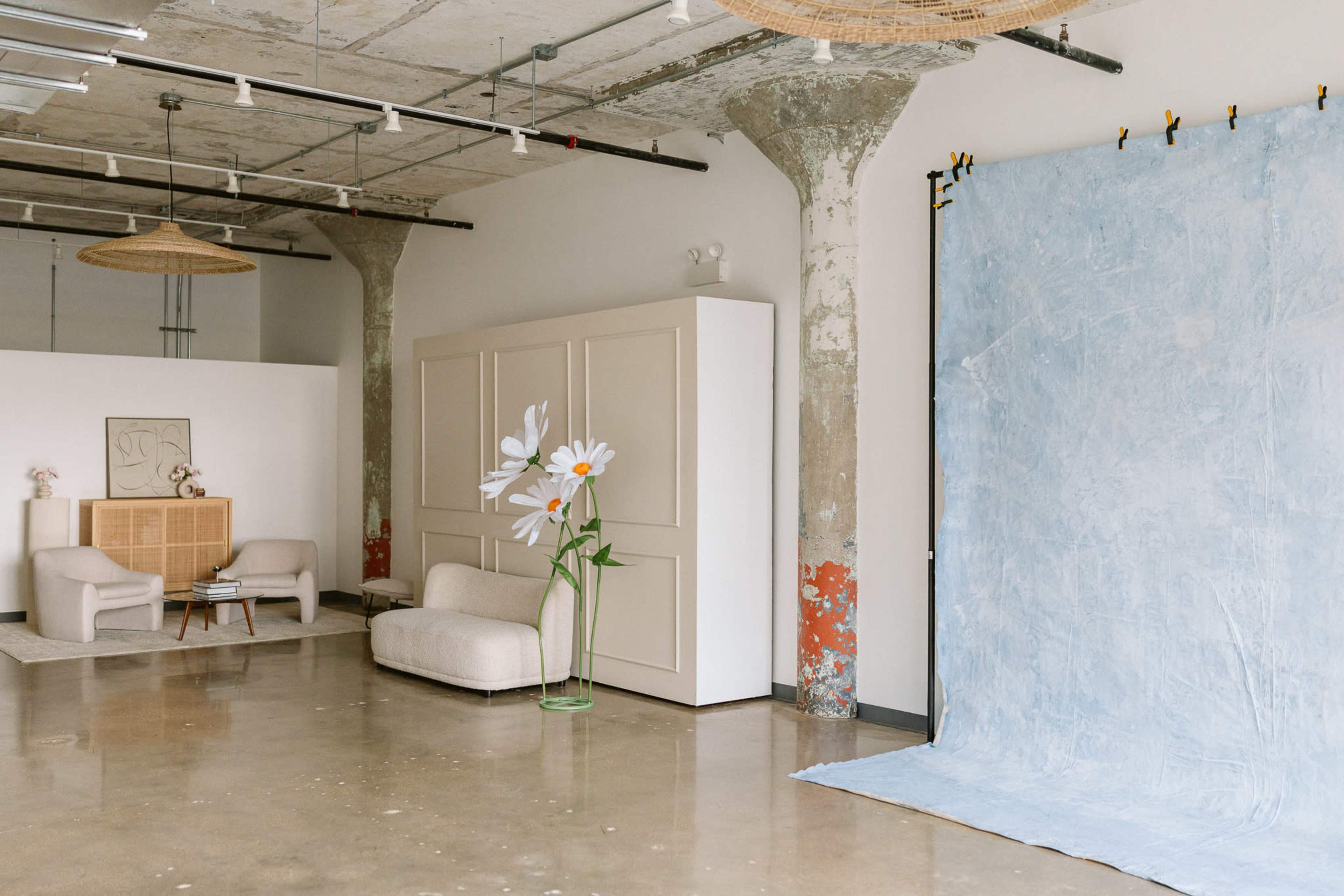 The image shows a spacious interior with concrete pillars, a light blue backdrop on one side, and modern furniture including a beige sofa and armchairs.