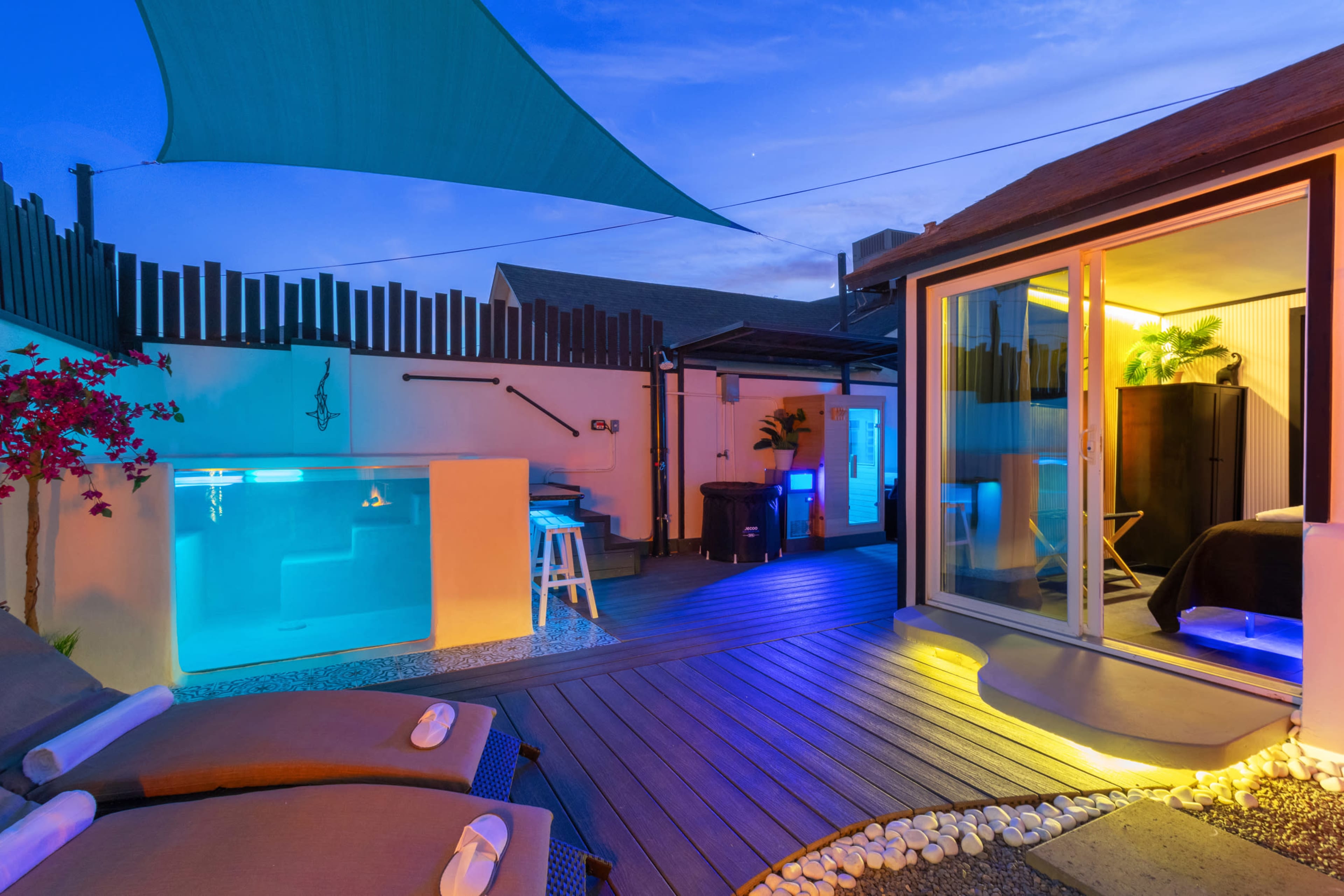 The image shows a modern outdoor patio area featuring a small illuminated pool, lounge chairs, and a glass door leading to an interior space, all set under a dusk sky.