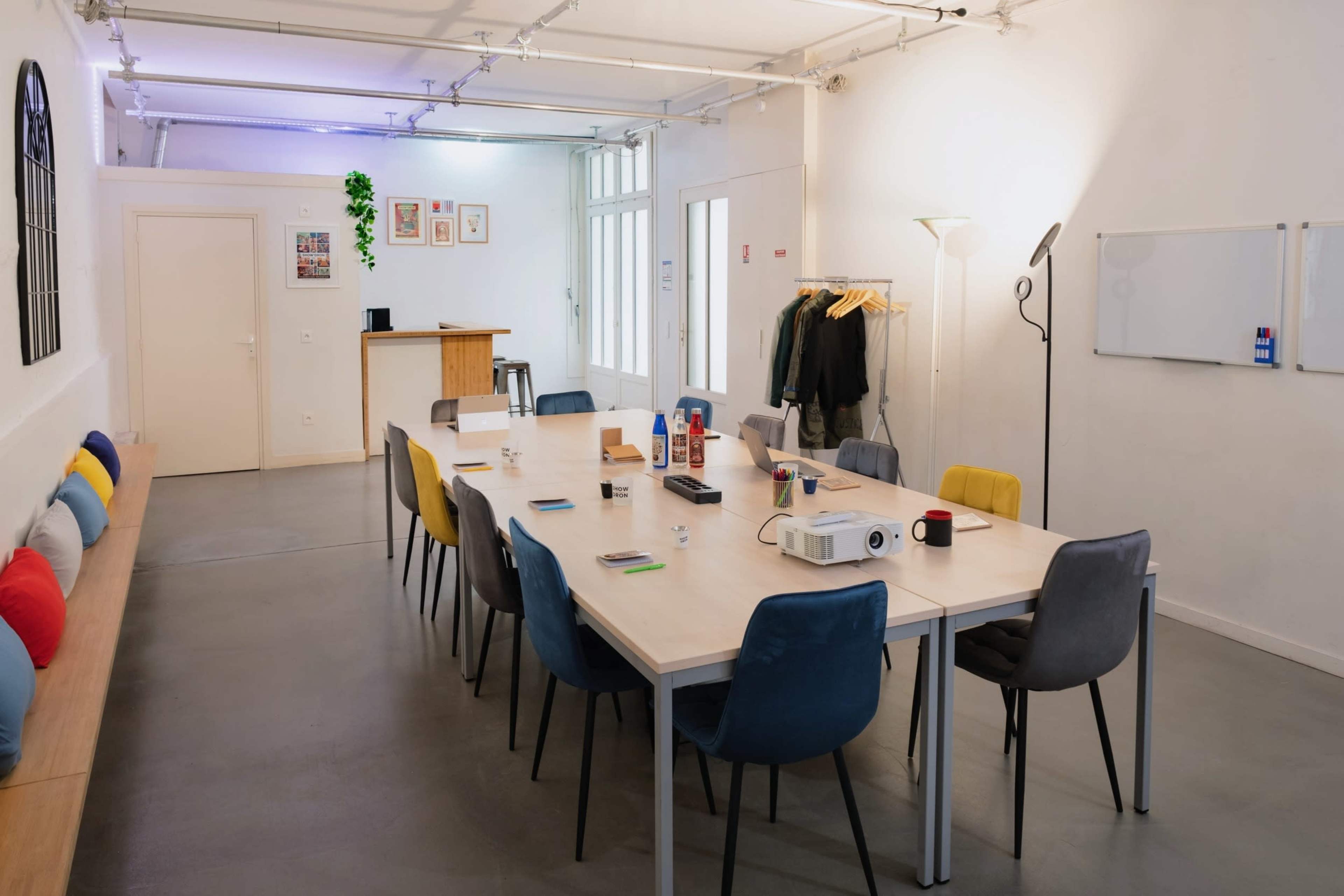 A modern, well-lit meeting room features a long table with colorful chairs, a projector, and a bar area, along with wall-mounted and floor lamps.