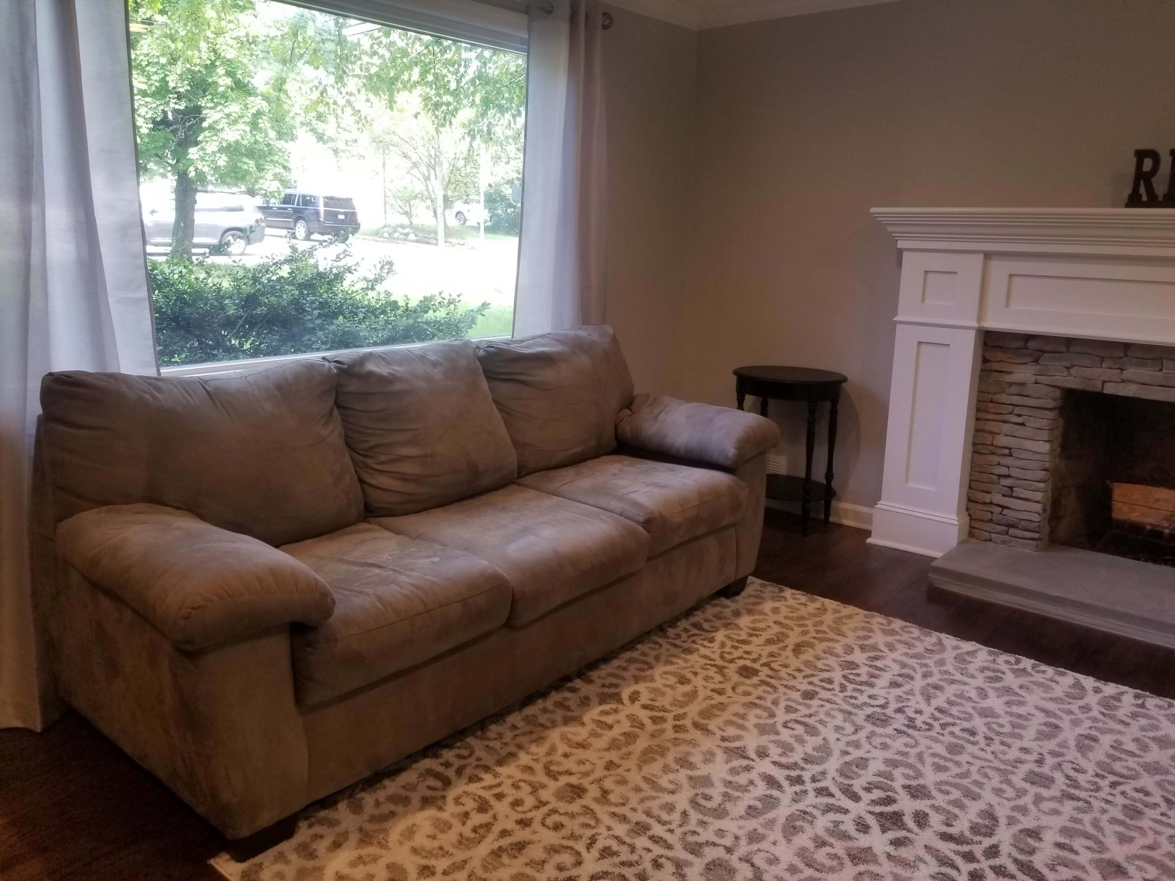 A tan sofa sits in a room beside a stone fireplace, with a window showing a view of greenery outside.