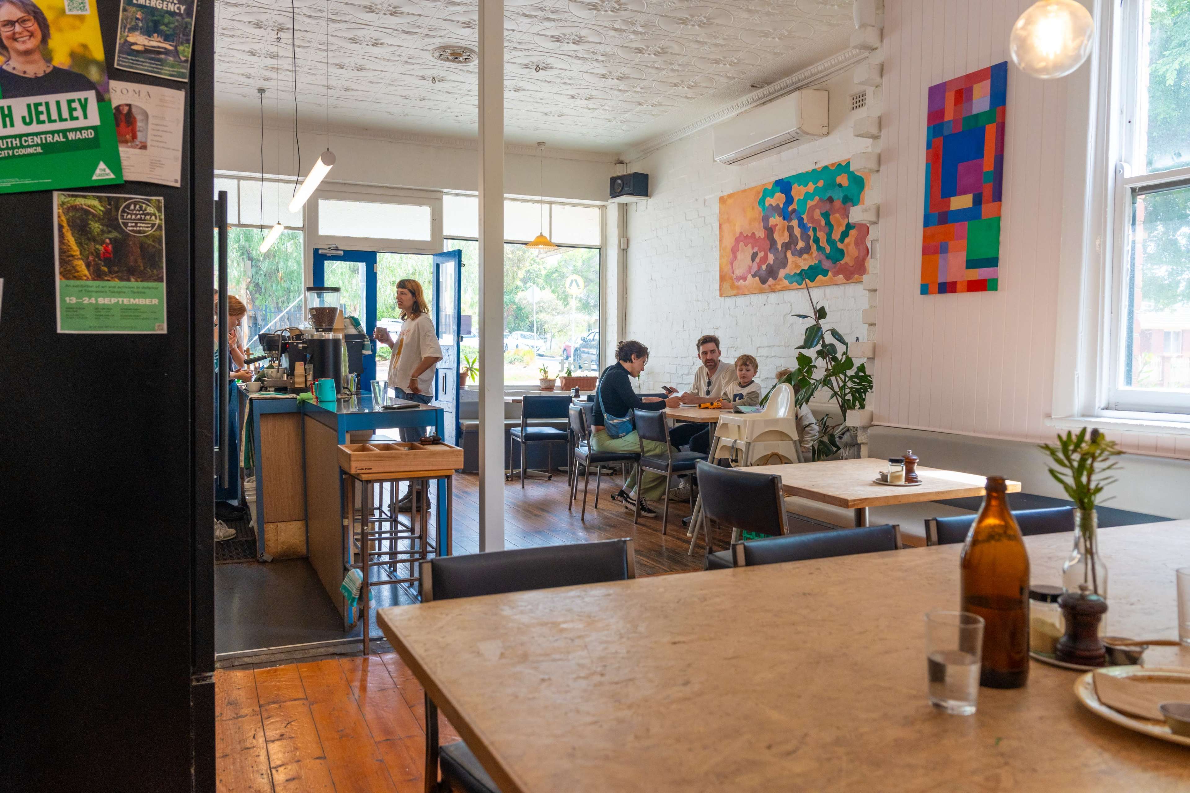 The image shows a casual café interior with wooden floors, colorful artwork on the walls, and patrons seated at tables.