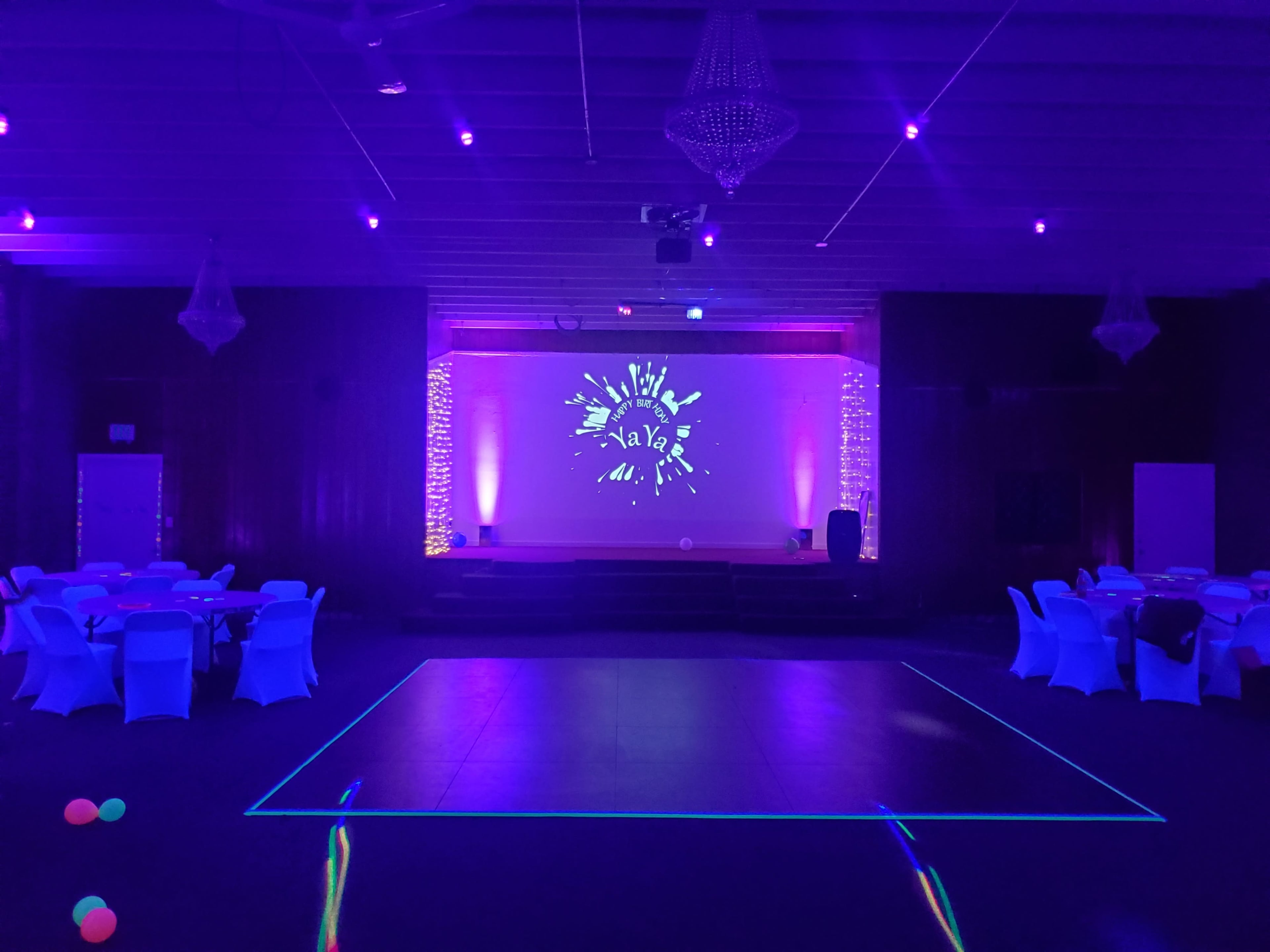 A dimly lit event space with purple lighting, featuring round tables covered in white cloths and a stage displaying a graphic with the word "ava."