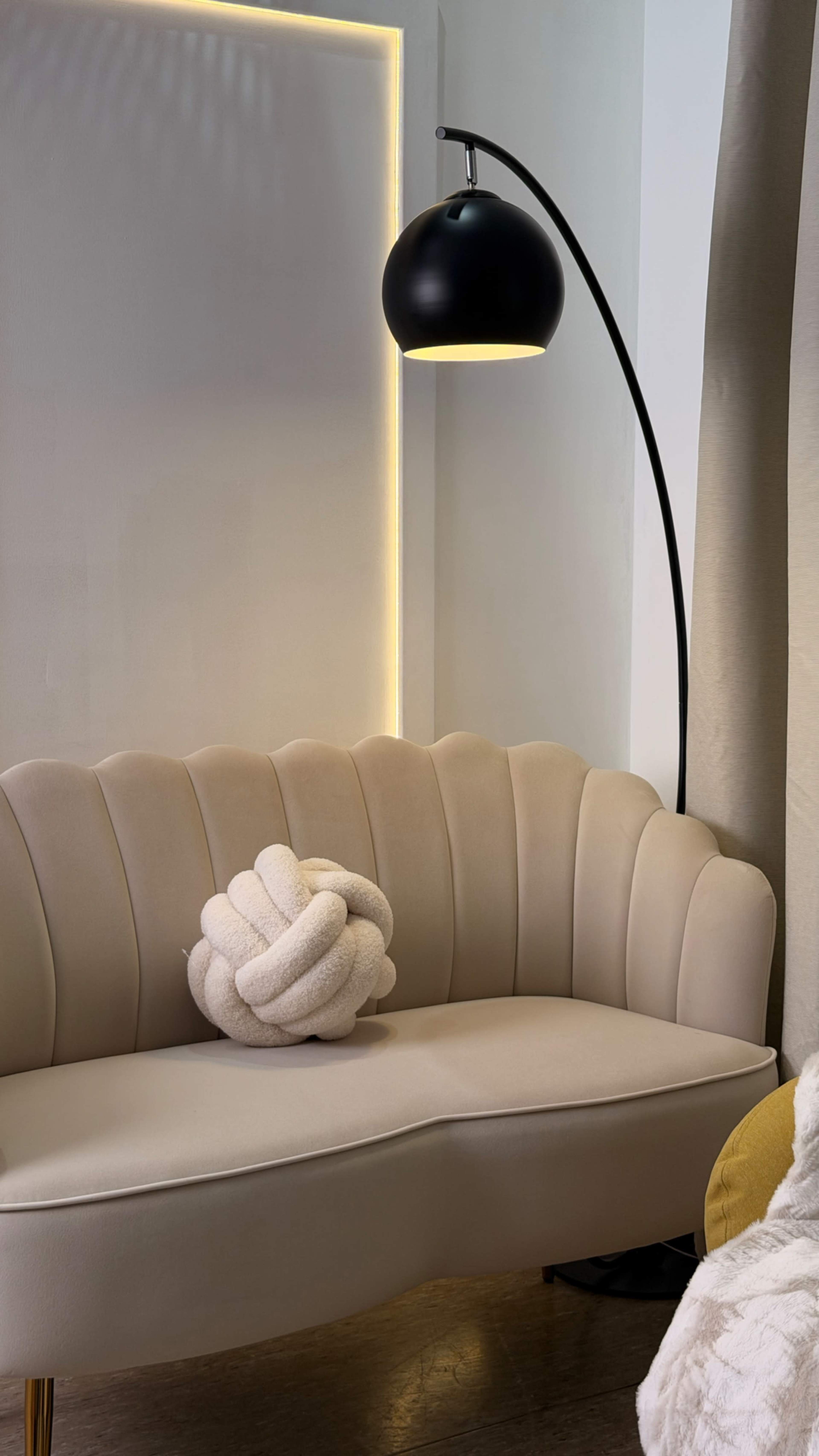 A curved beige sofa with a decorative knot-shaped cushion and a black arc floor lamp is positioned against a softly lit wall.
