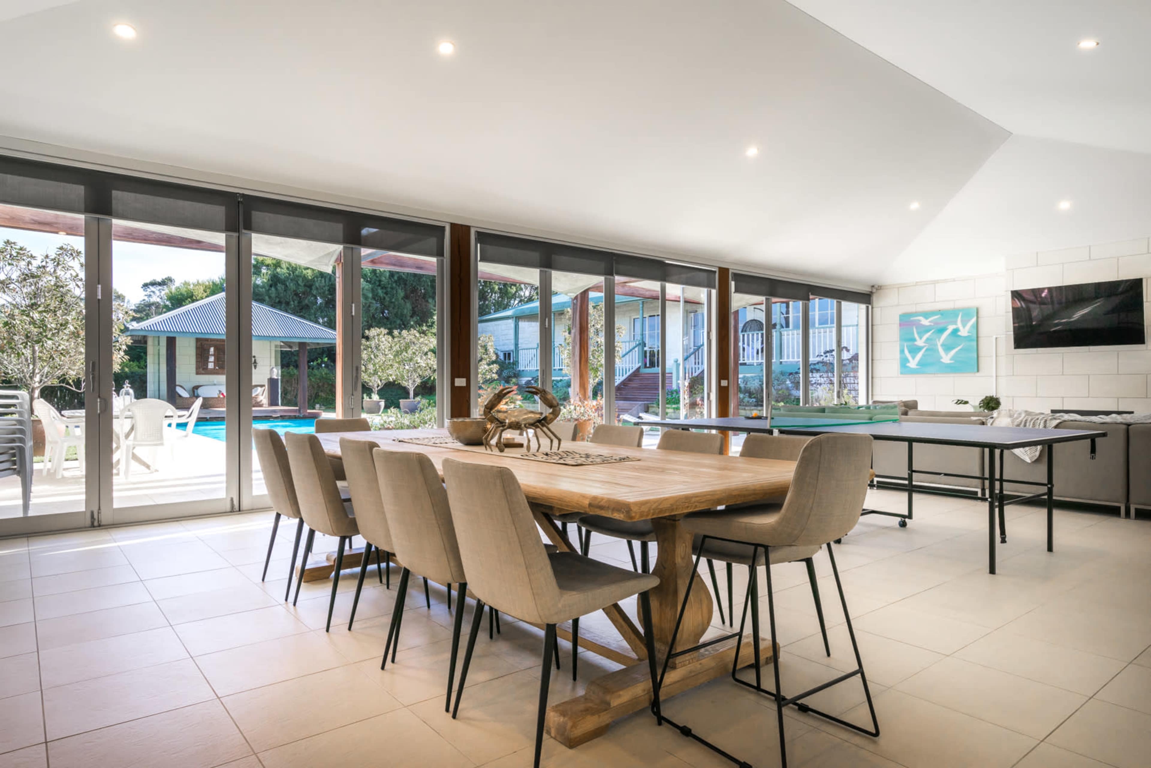 A spacious dining area features a long wooden table with upholstered chairs, large windows overlooking a backyard with a pool, and a modern decor.