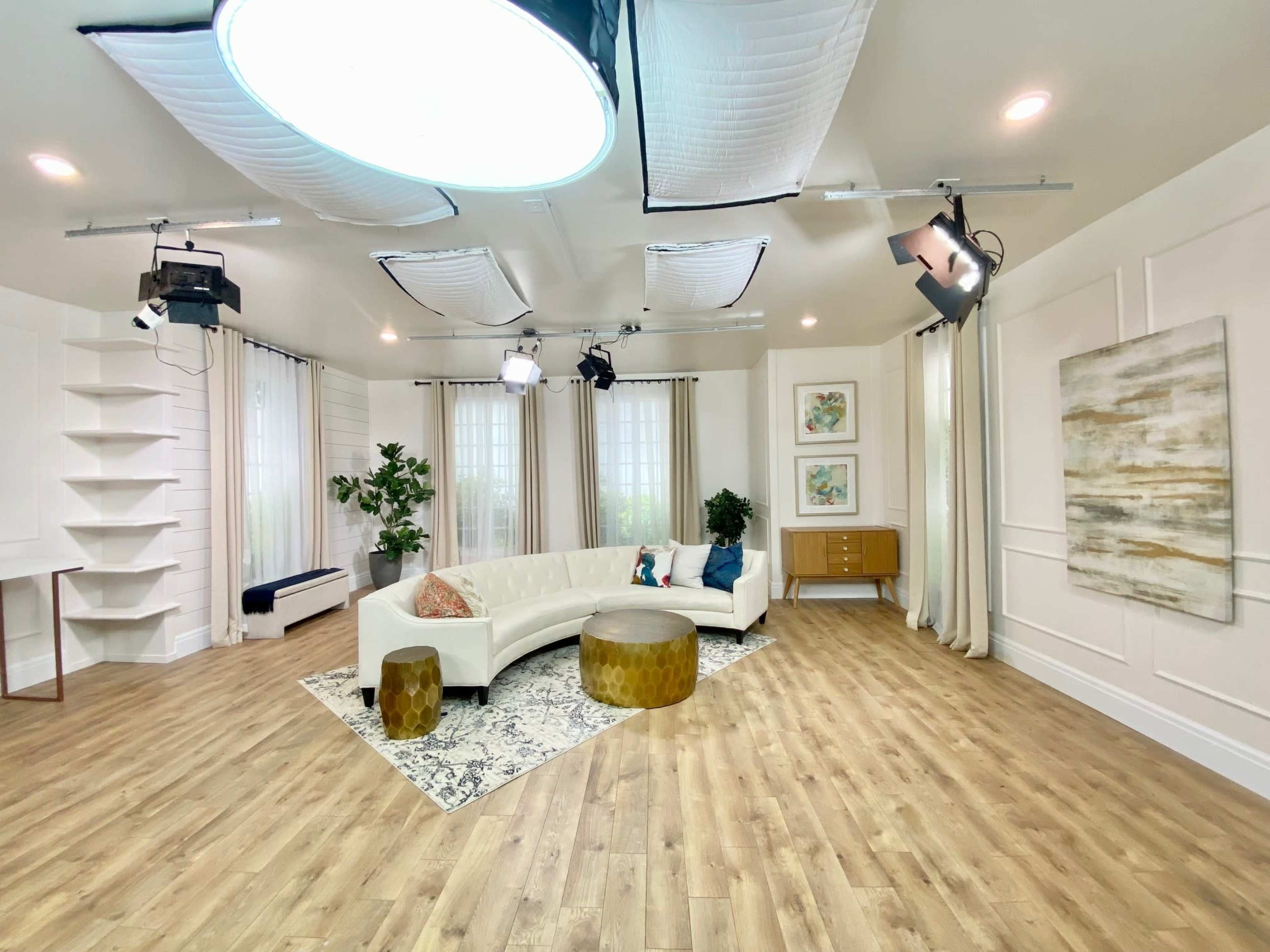 A well-lit studio space featuring a curved white sofa, a round gold table, and decorative plants against a backdrop of wall art and curtains.