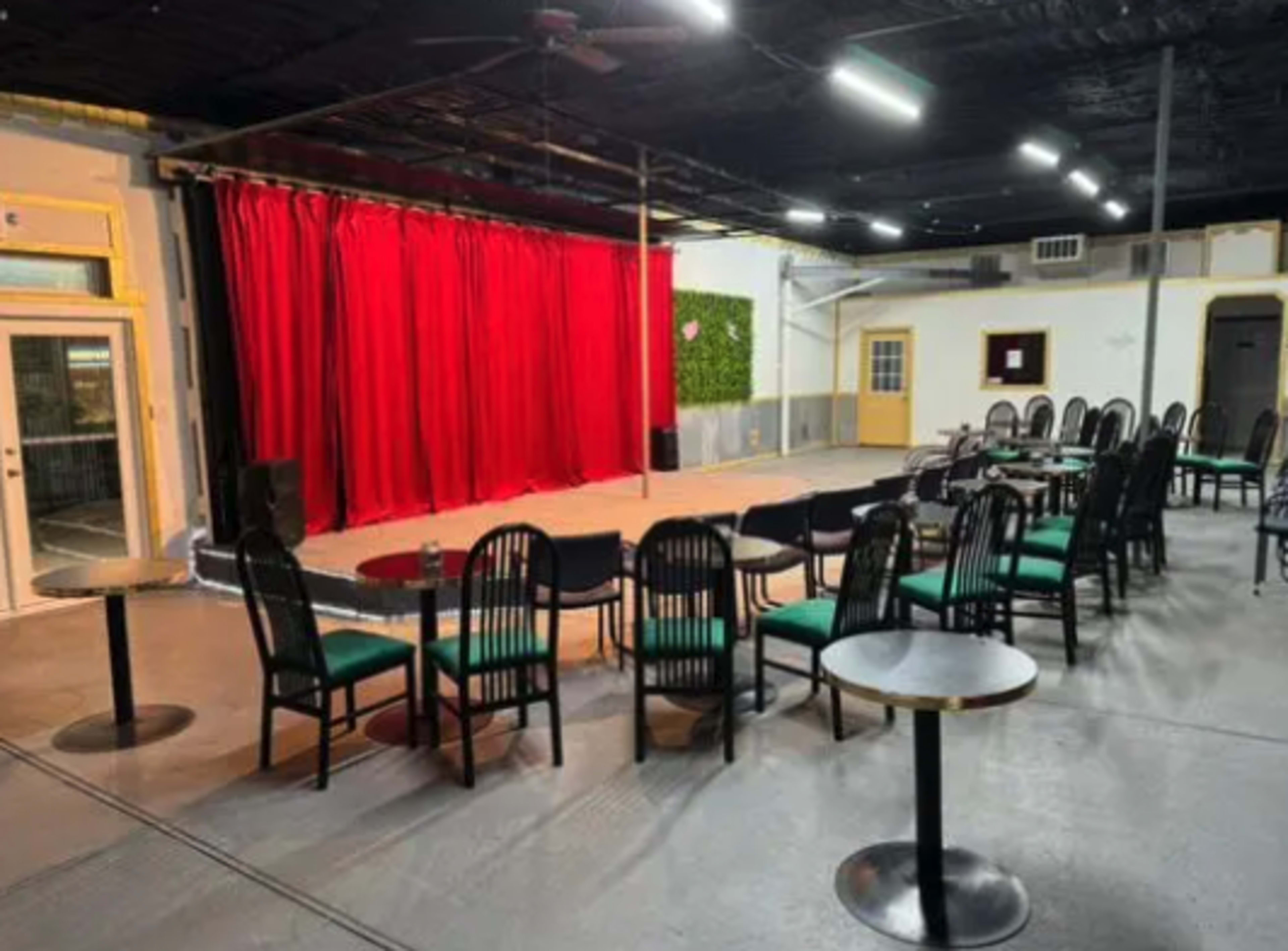 A performance venue with a red curtain, a stage area, and rows of black chairs arranged around small tables.