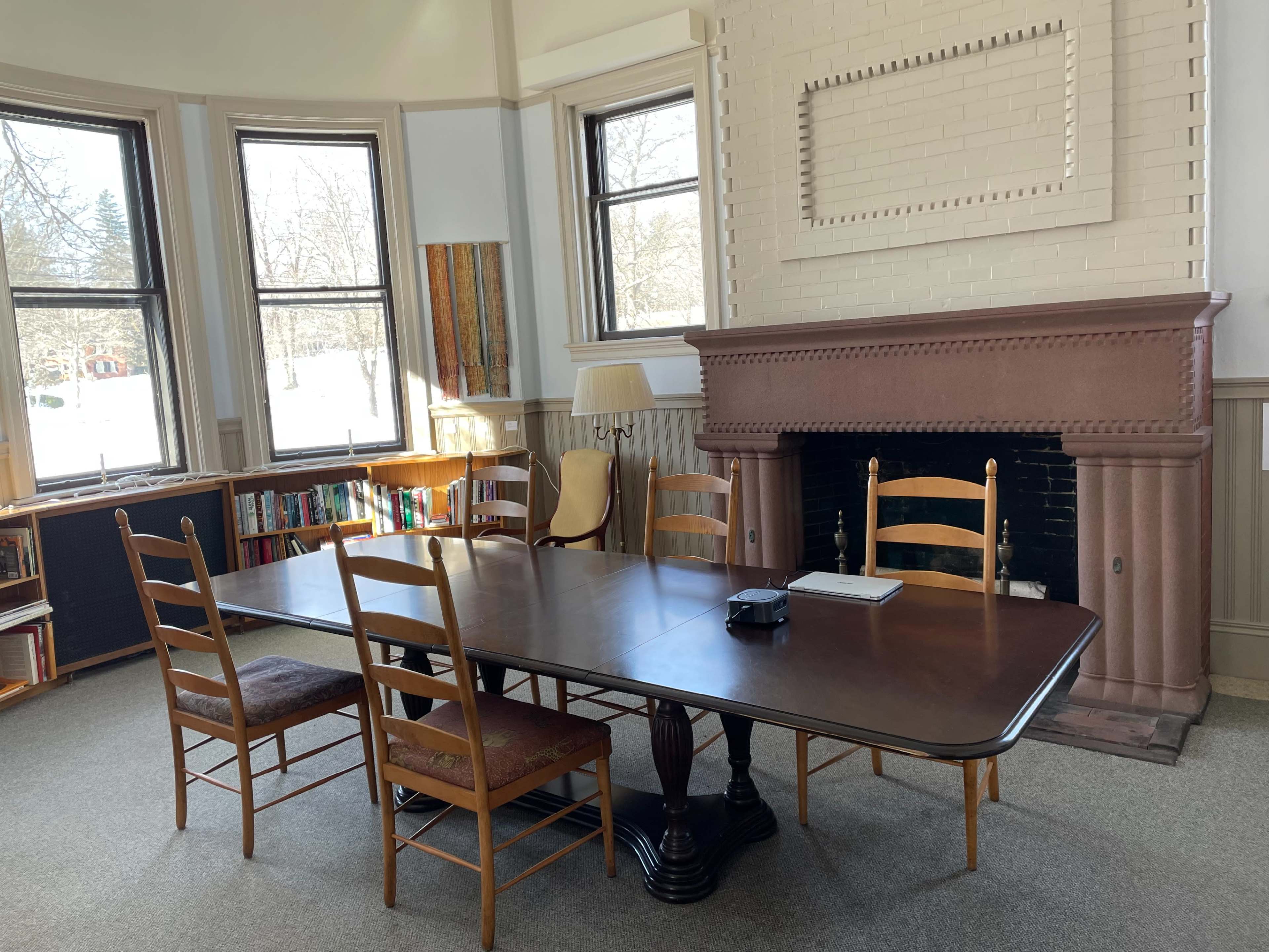 A rectangular wooden table with several chairs is positioned in a bright room featuring large windows and a brick fireplace.