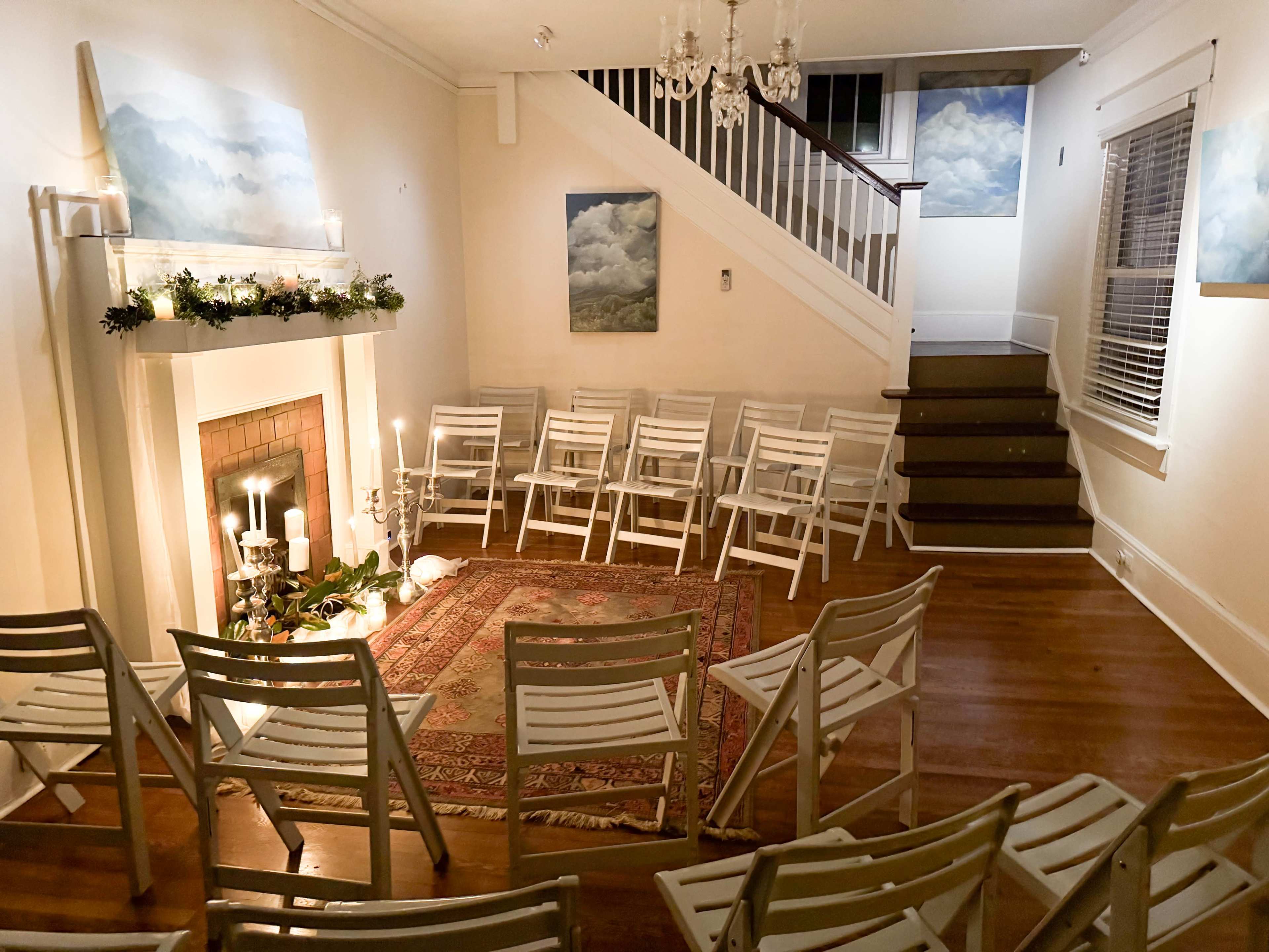 A neatly arranged room with white chairs set up around a decorative area featuring a rug and a fireplace, with paintings of clouds on the walls.