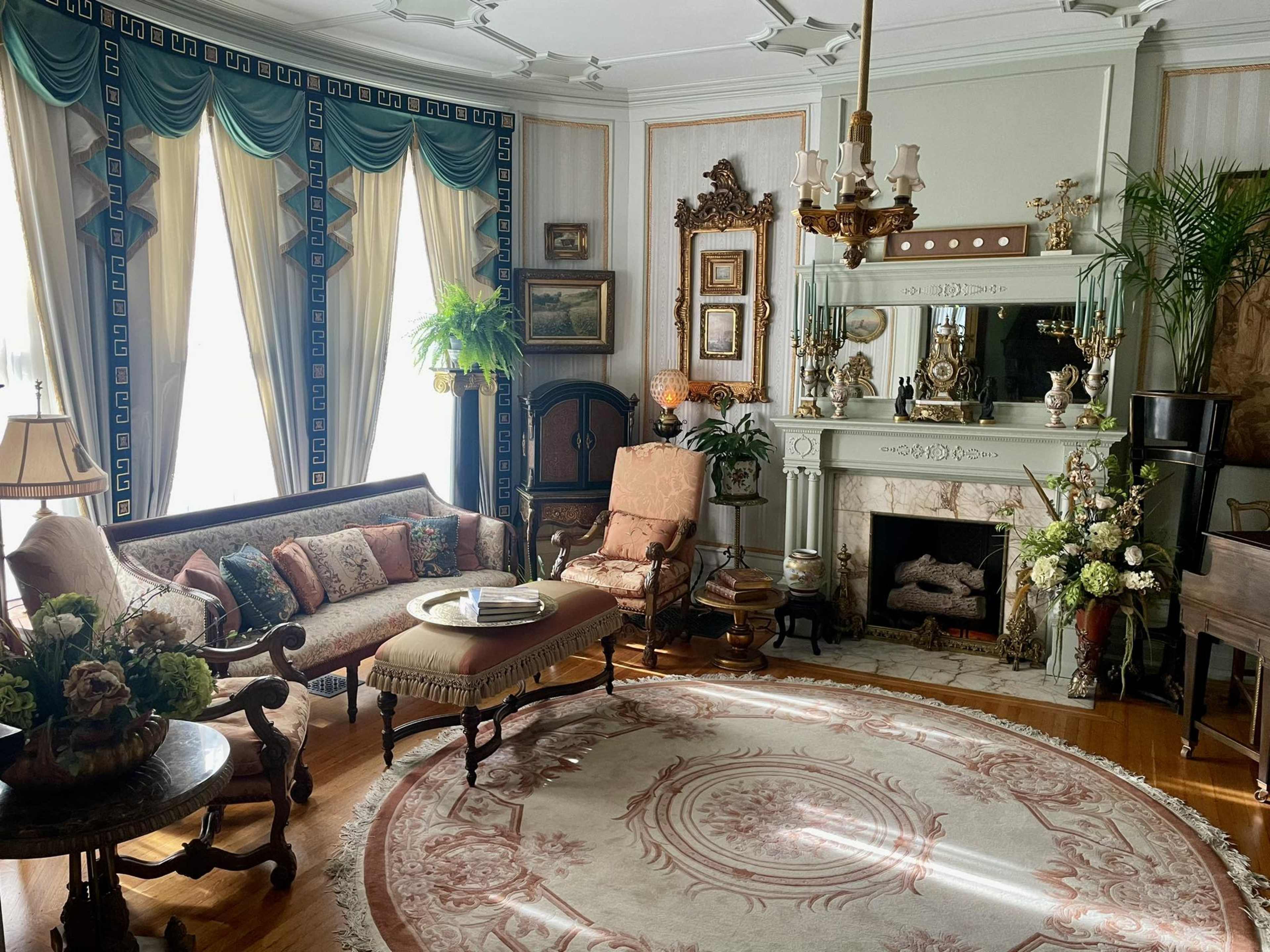 The image depicts a vintage living room featuring ornate furniture, a decorative fireplace, and elaborate drapery, set against a patterned rug.