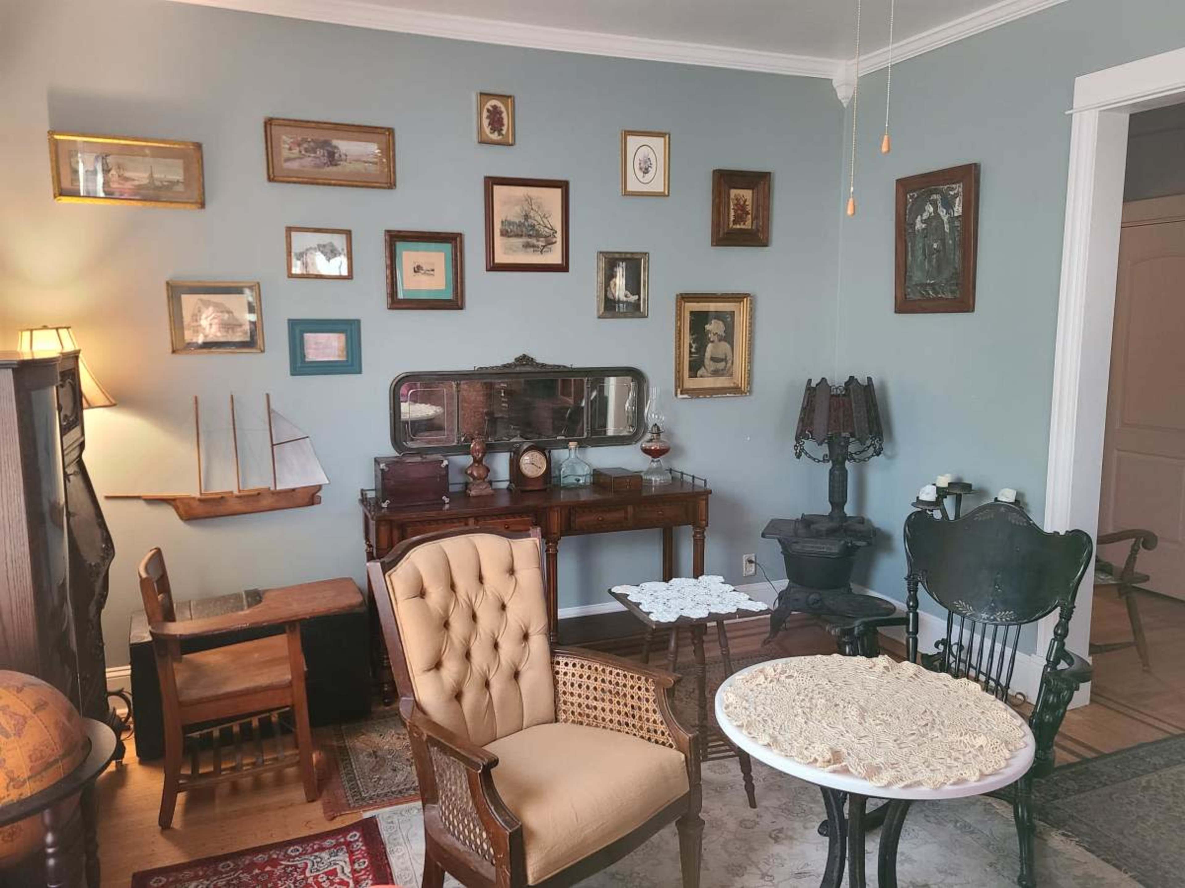 The room features vintage furniture, a small sailboat model, and a wall adorned with various framed artworks and photographs.