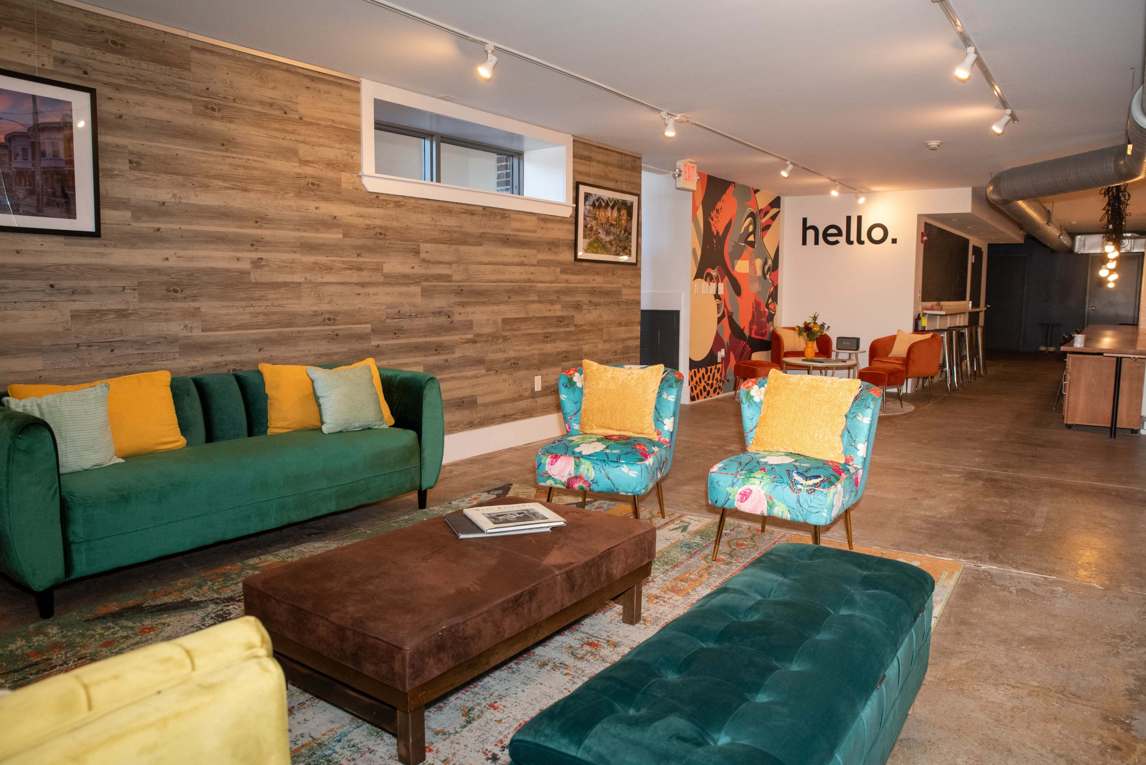The image shows a modern, stylish lounge area with green sofas, floral chairs, a brown coffee table, and a vibrant mural featuring the word "hello" on the wall.