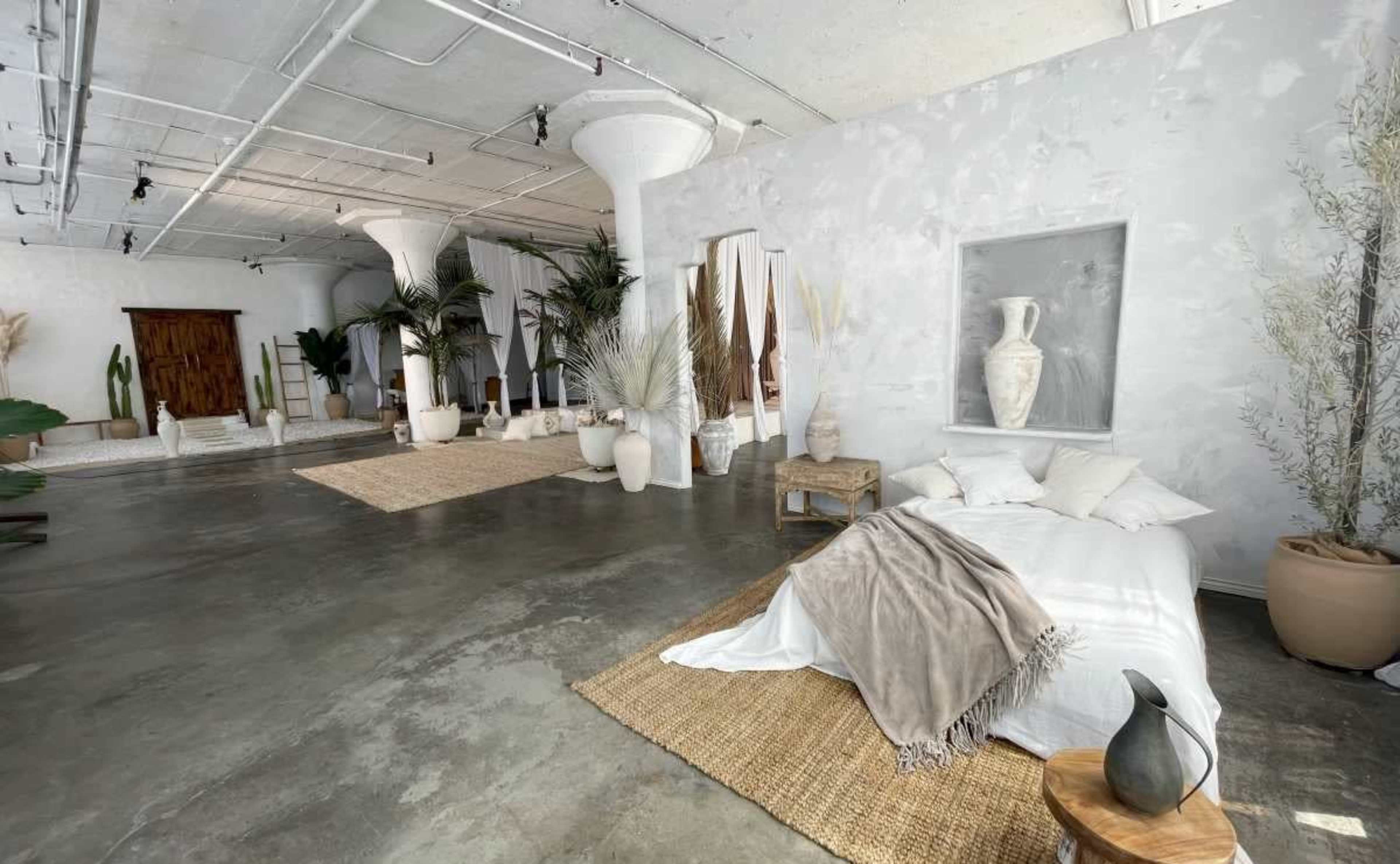 The image shows a minimalist studio space with a bed made of white linens, surrounded by various decorative plants and pottery on a polished concrete floor.