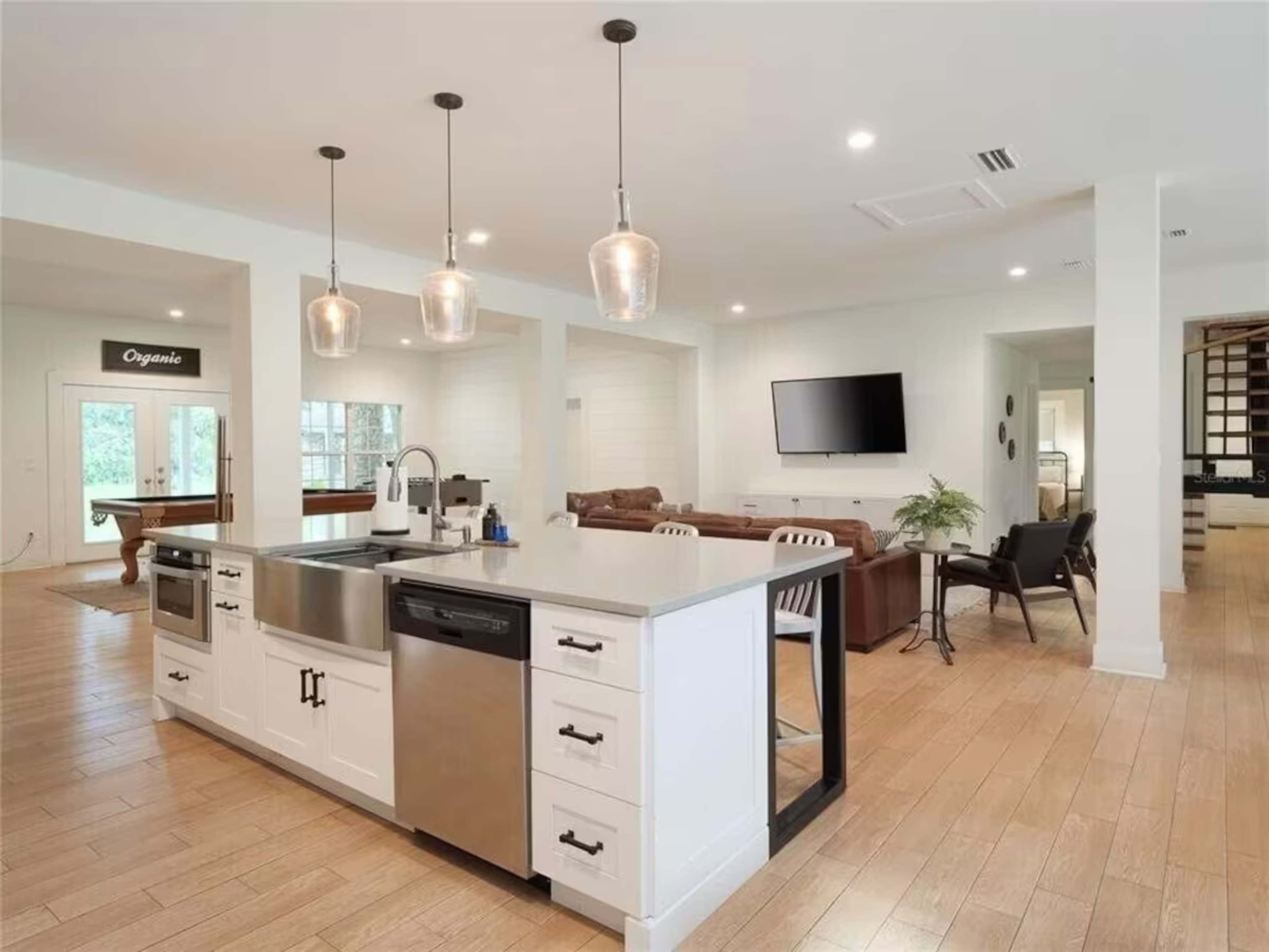 A modern open-concept kitchen features a large island with a sink, stainless steel appliances, and pendant lighting, leading into a spacious living area with a TV and seating.