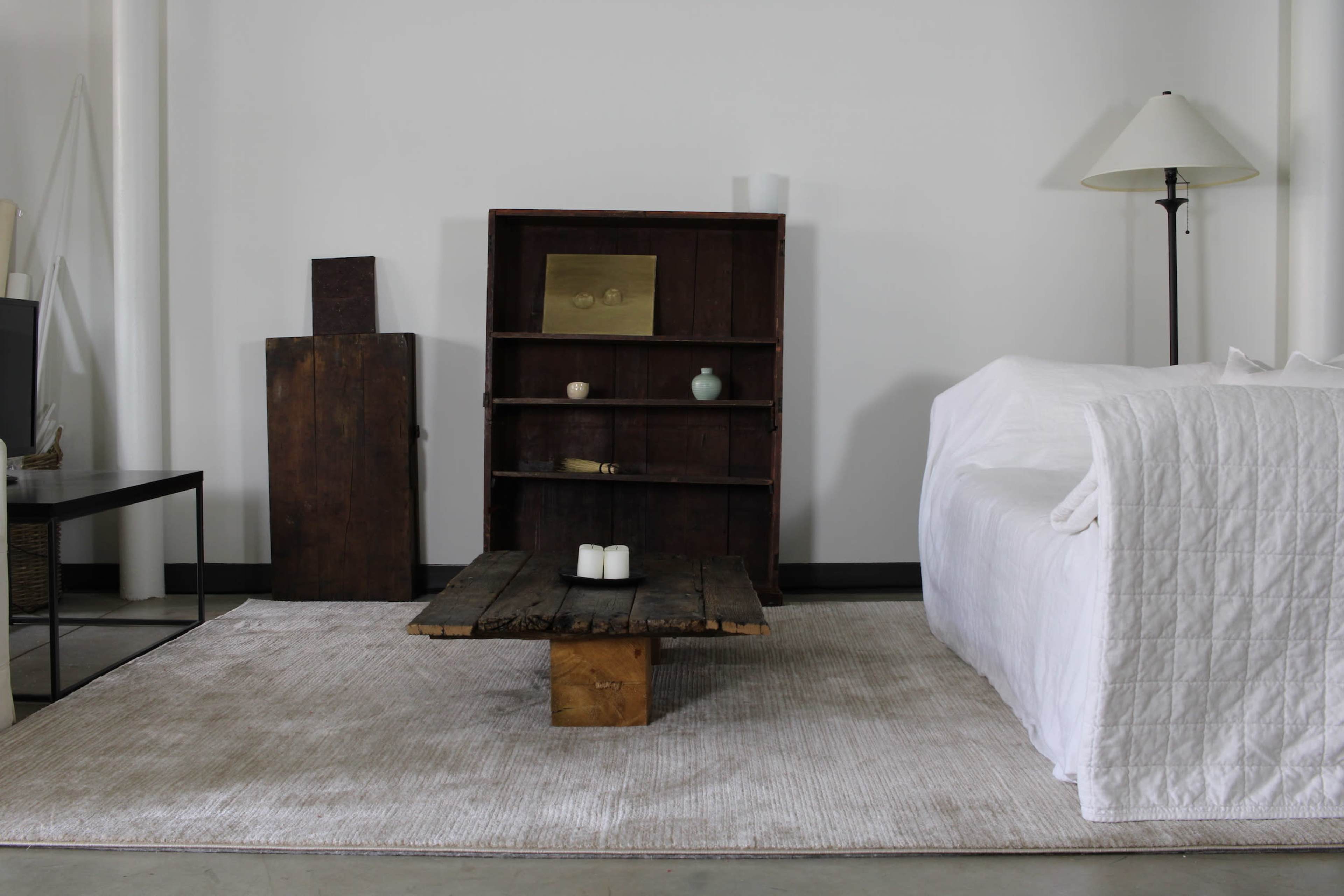 A minimalist living room with a wooden coffee table, a white sofa covered with a blanket, and a dark wooden bookshelf against the wall.