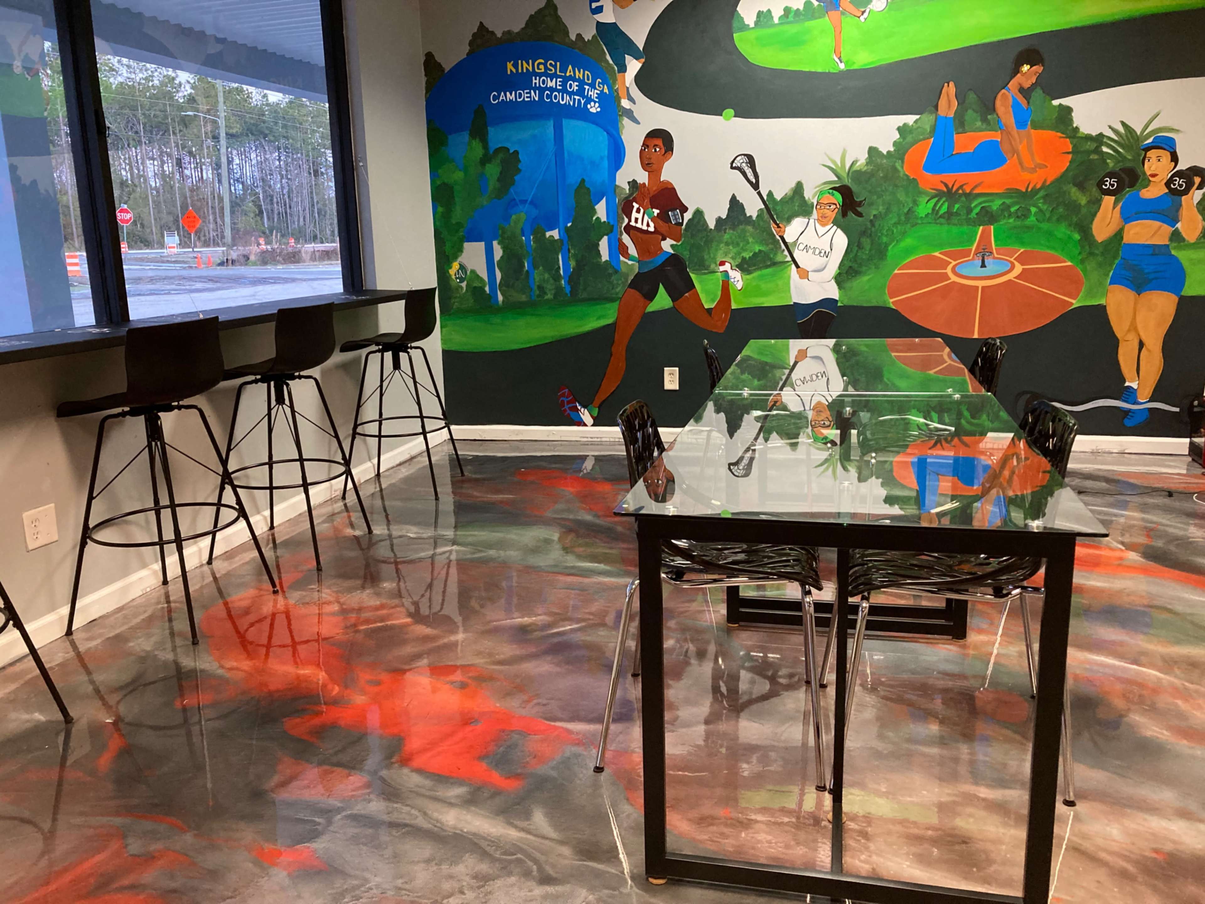 A modern room with a glass table and black chairs, featuring a colorful mural depicting athletes and a polished floor with a swirling pattern.