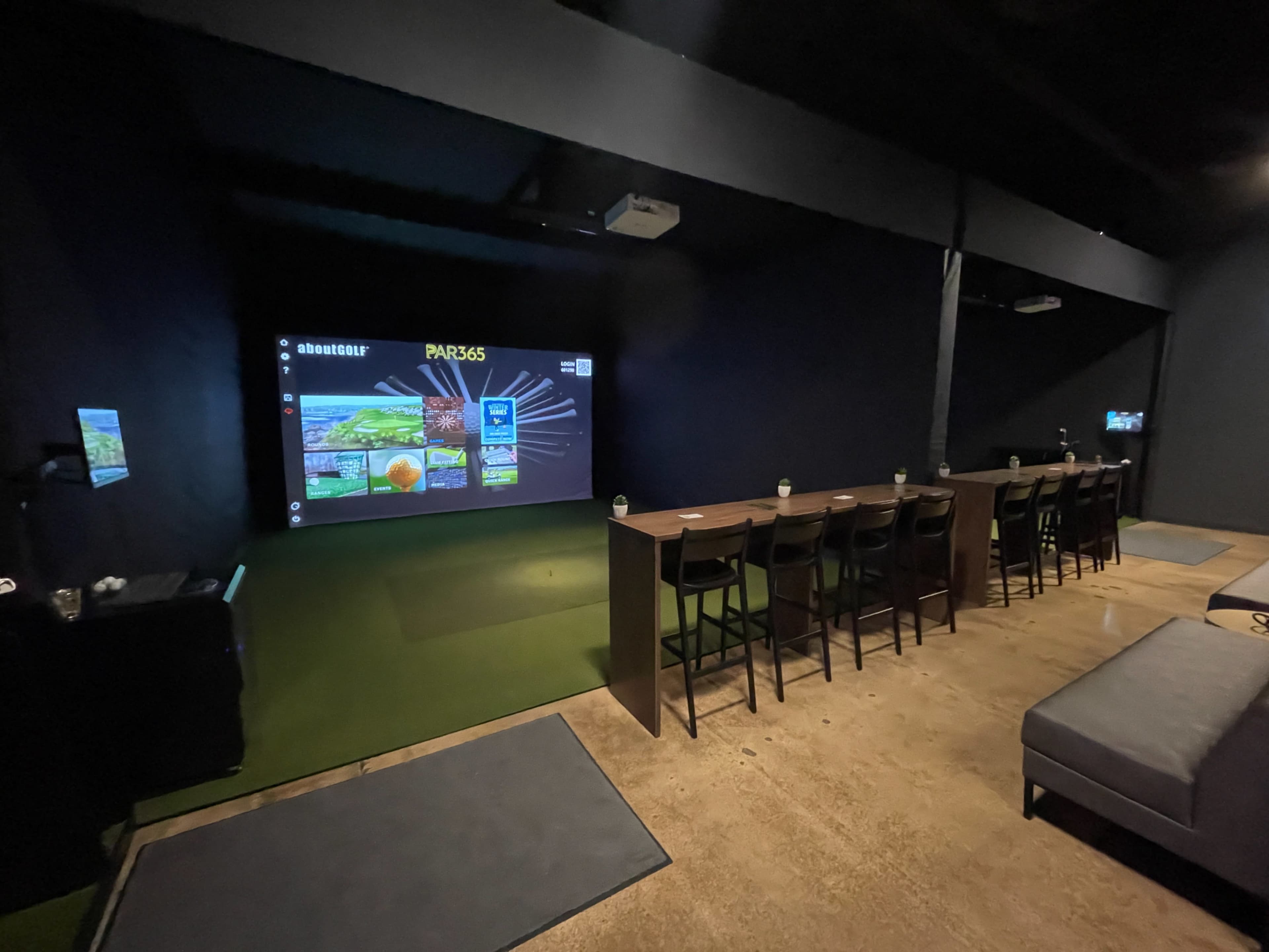The image shows a dimly lit indoor space with a long table featuring high chairs, a projector screen displaying a golf simulation, and a sofa along one wall.