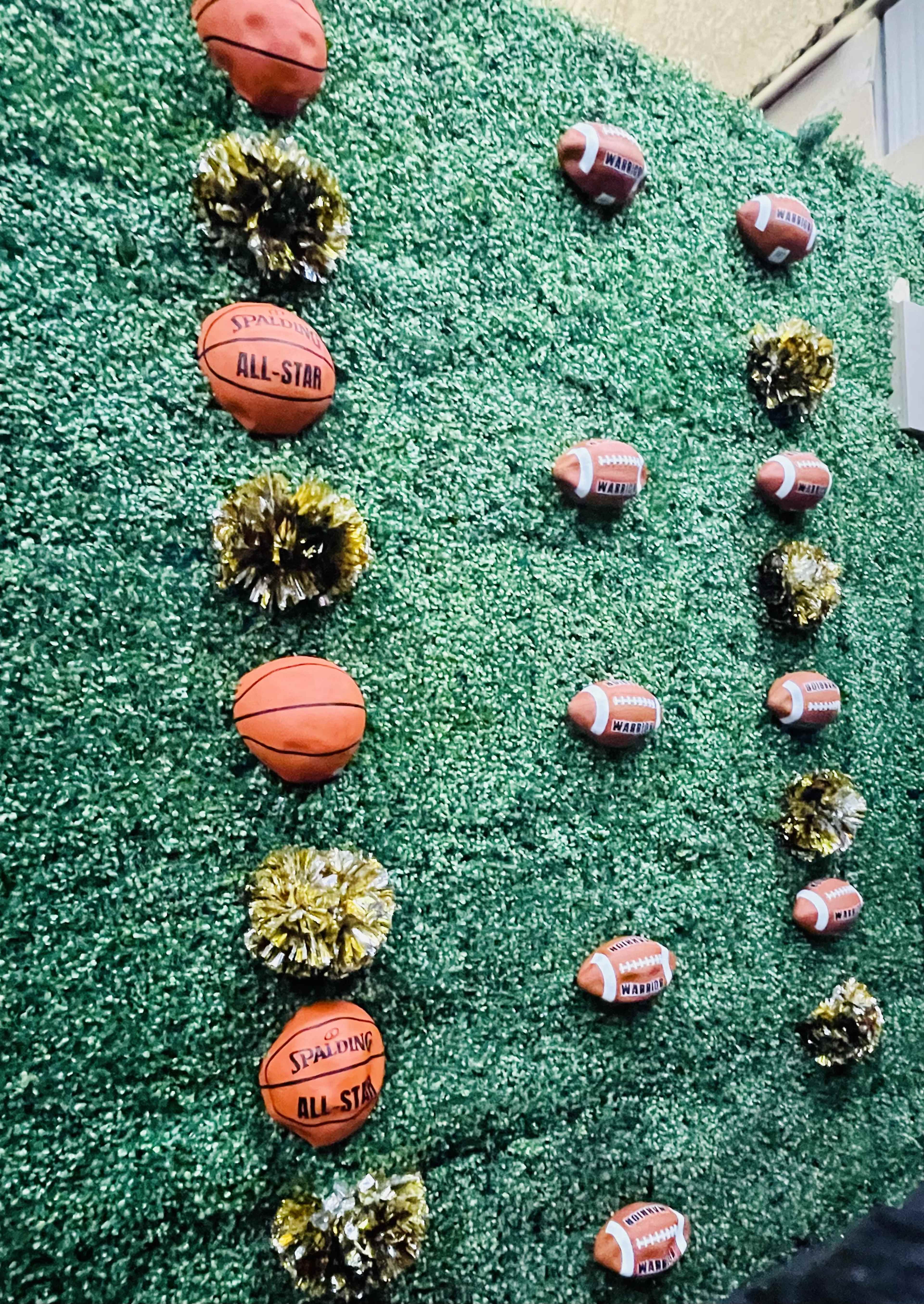 The image shows a green wall decorated with basketballs and footballs alongside yellow and gold pom-poms.