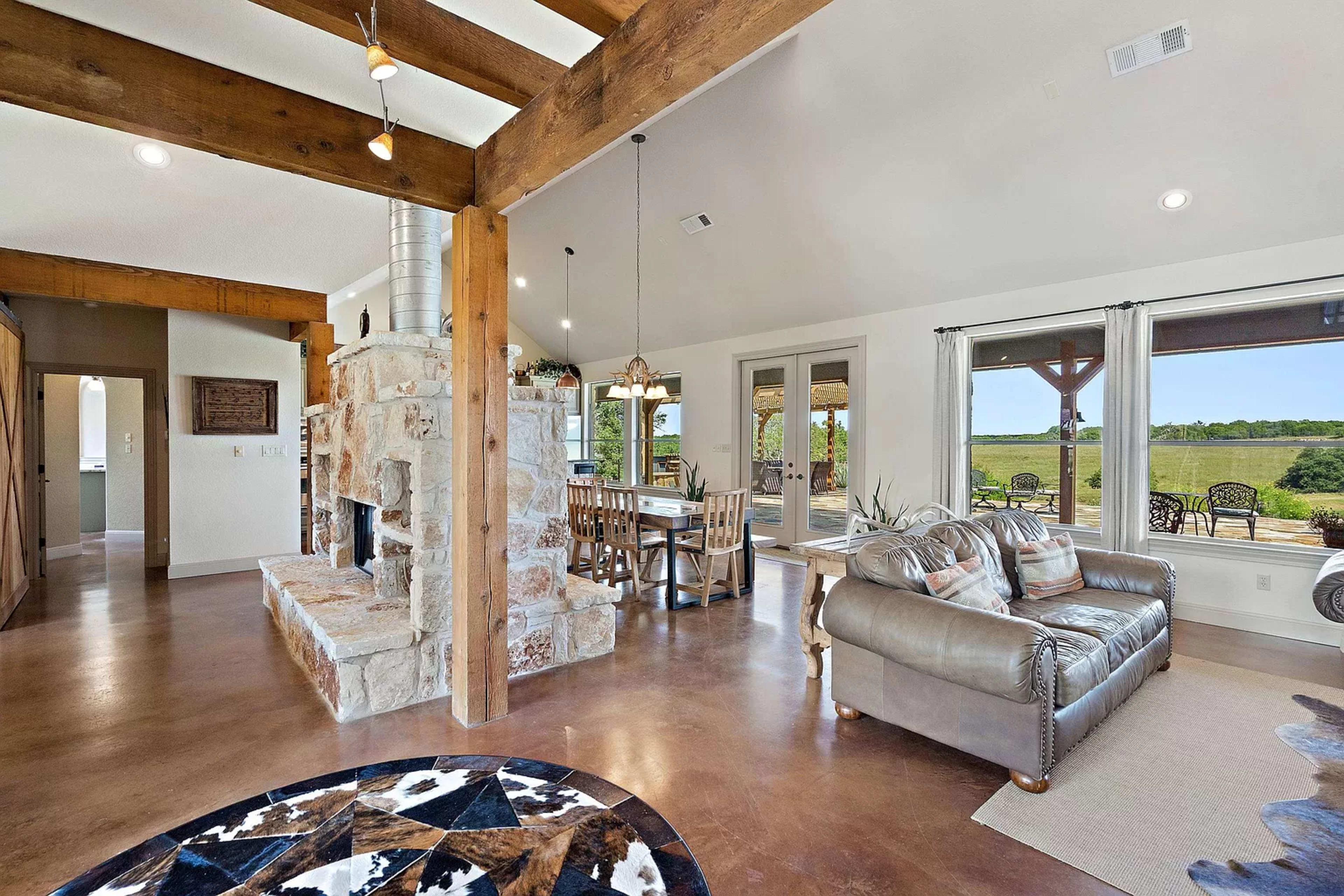 The image shows a spacious living area with a stone fireplace, rustic wooden beams, and large windows overlooking a green outdoor landscape.