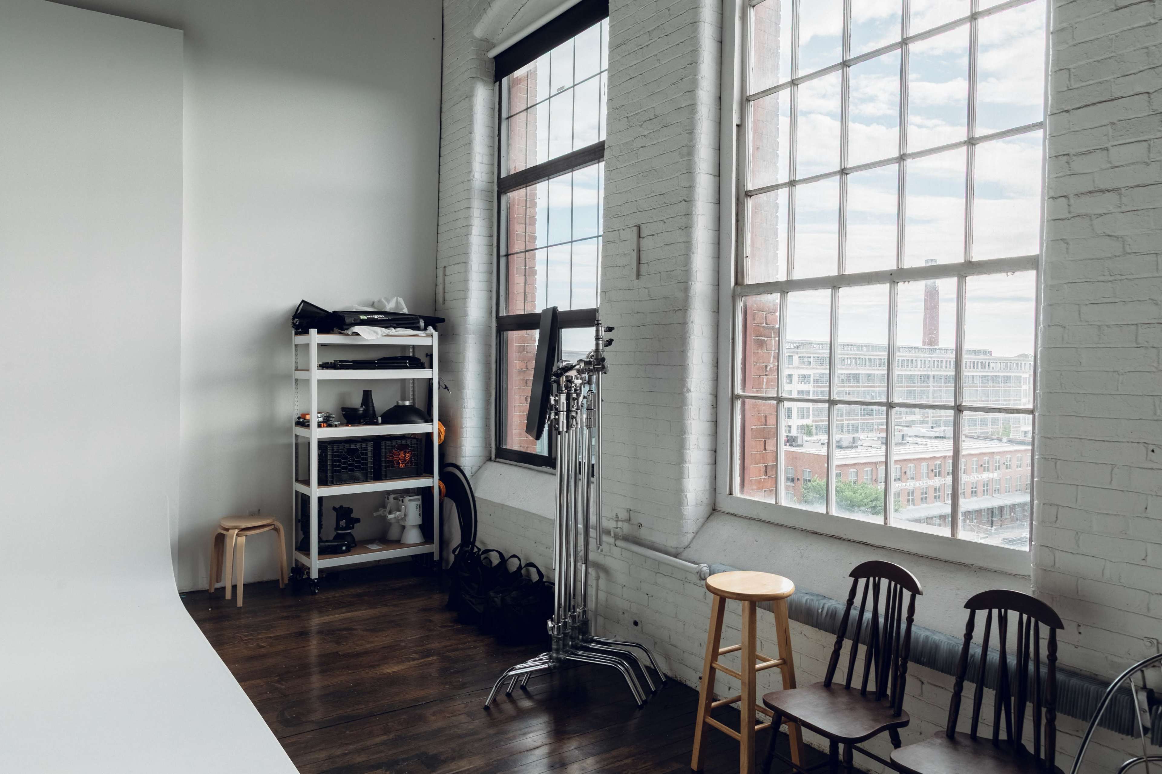A photography studio features a white backdrop, storage shelves with equipment, and several wooden chairs beside large windows.
