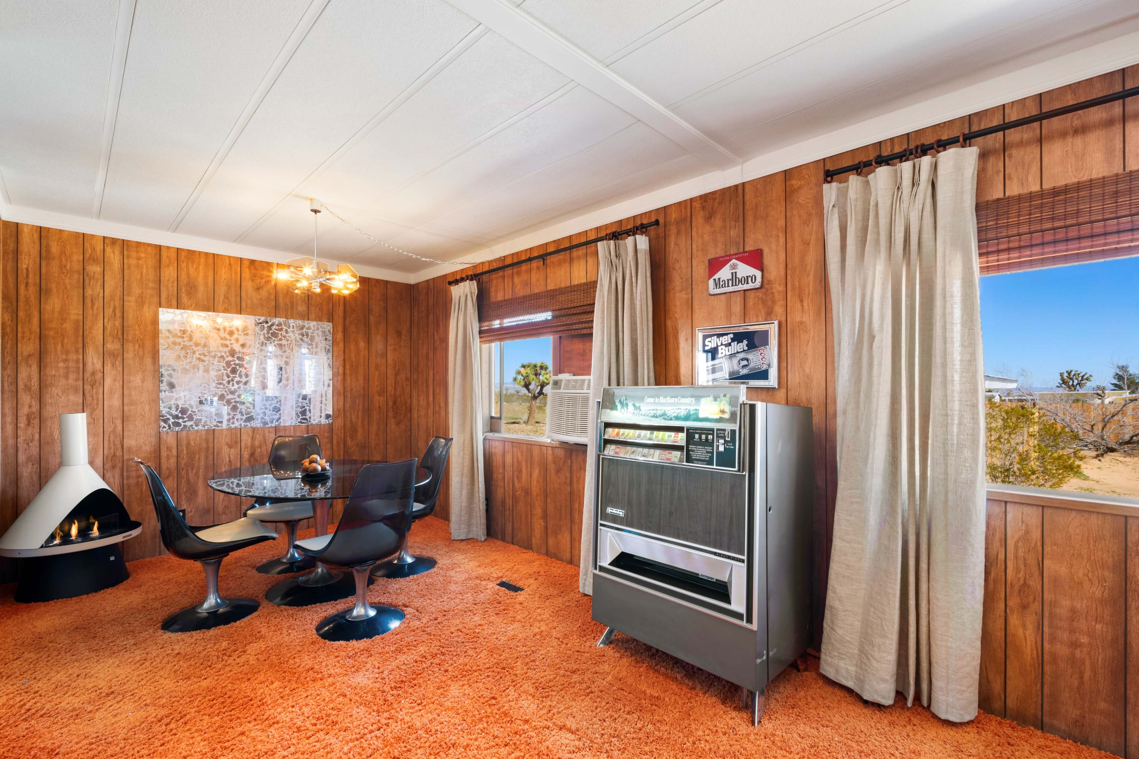 The image shows a retro-styled living room with wooden paneling, a glass dining table, and a vintage beverage cooler against a backdrop of desert scenery visible through large windows.