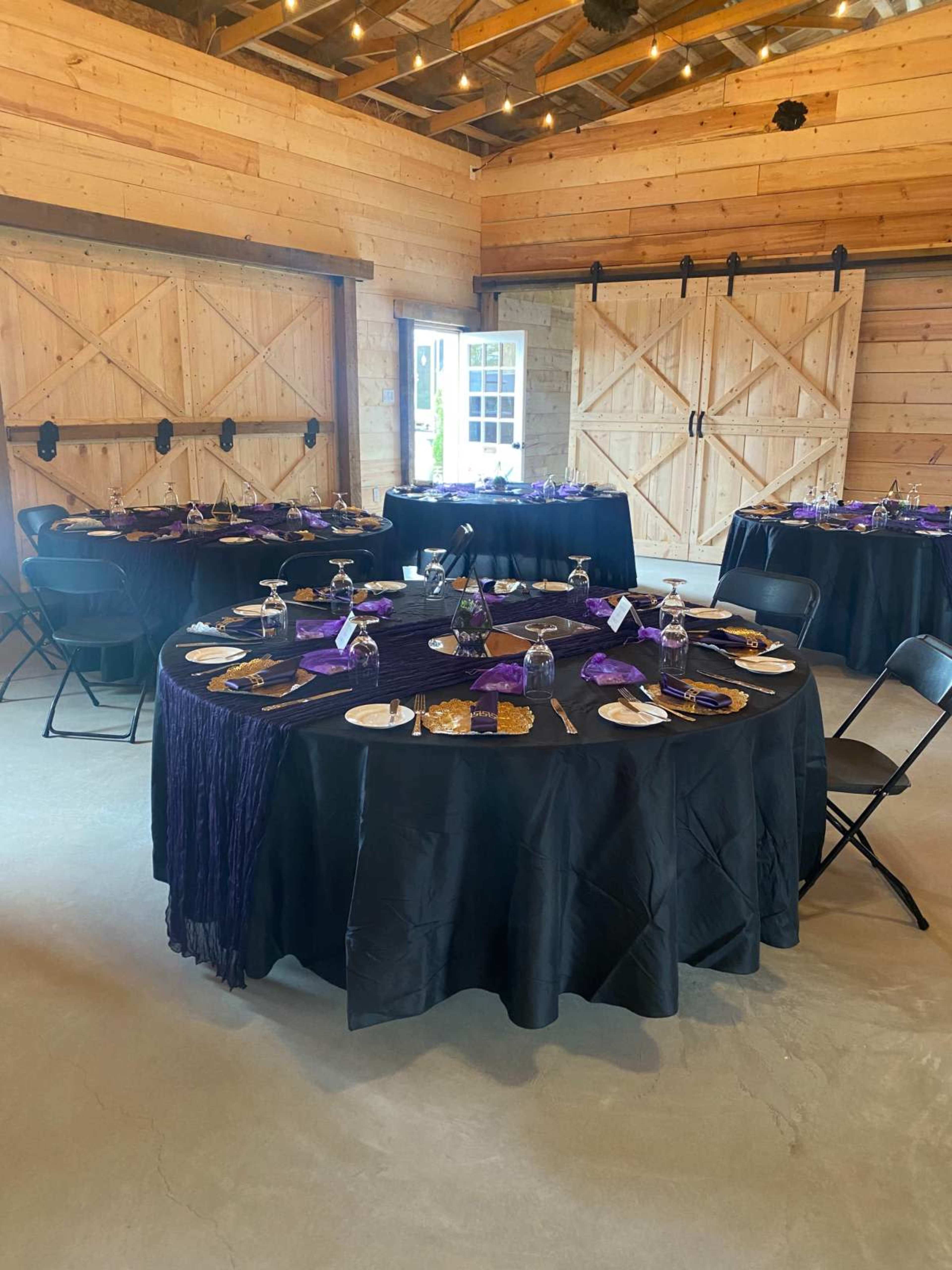 The image shows a spacious venue with several round tables covered in black tablecloths, each set with purple decor, glassware, and dishes, against a backdrop of wooden walls and large doors.
