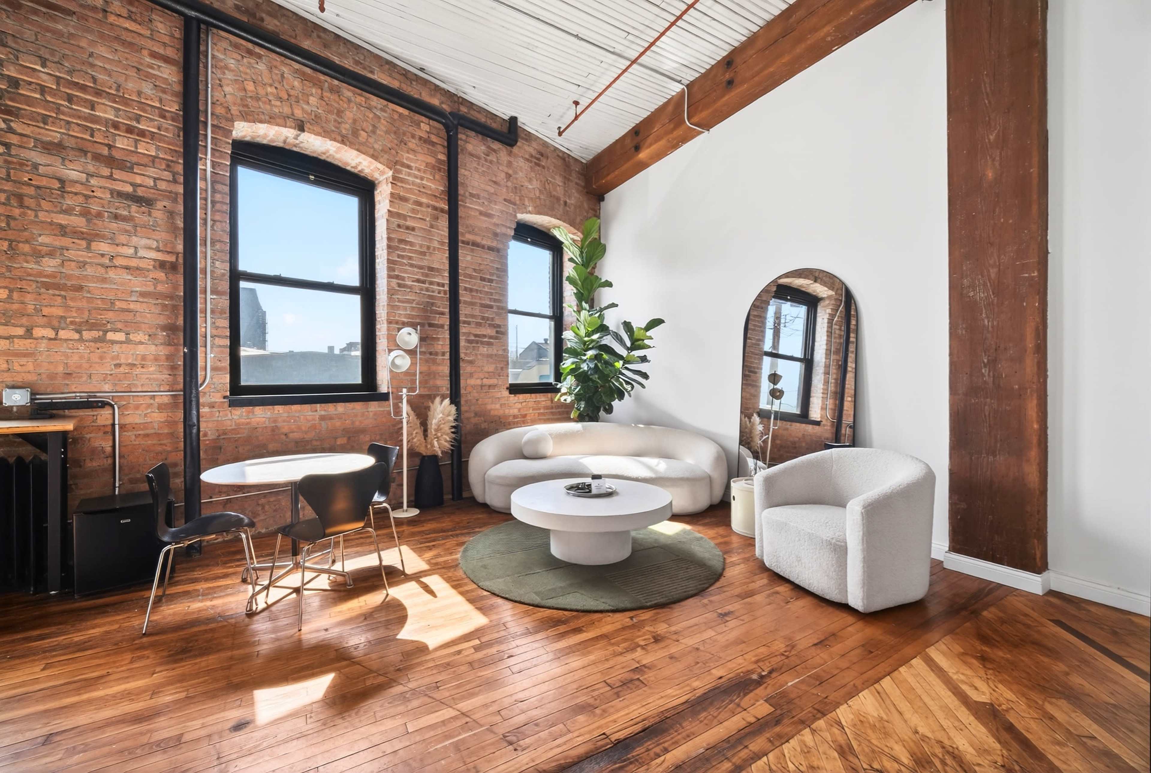 The image features a spacious interior with exposed brick walls, wooden beams, a round coffee table, and a white sofa, complemented by a plant and a chair.