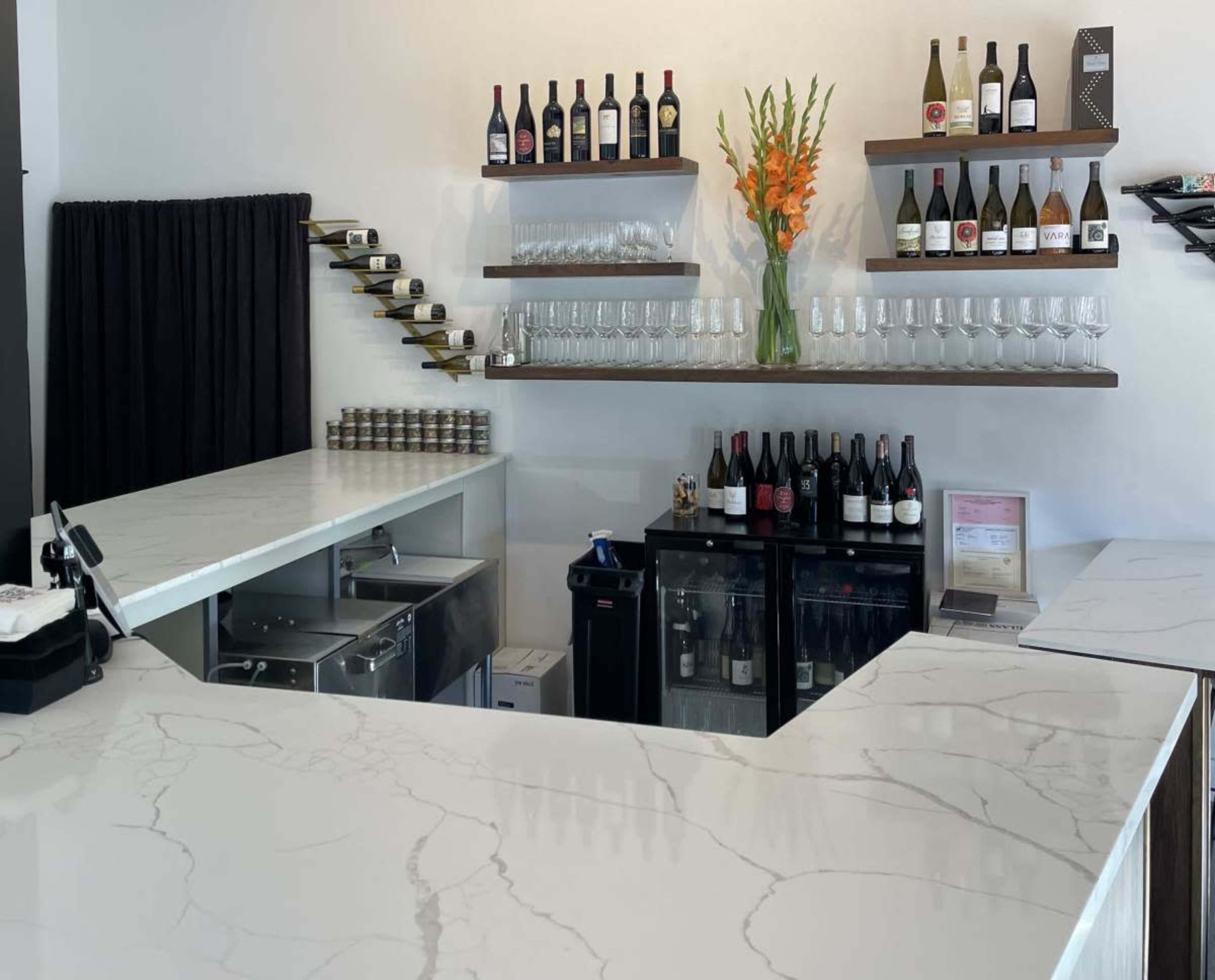 The image shows a modern bar area featuring a marble countertop, shelves with various bottles of wine, and a display of wine glasses.