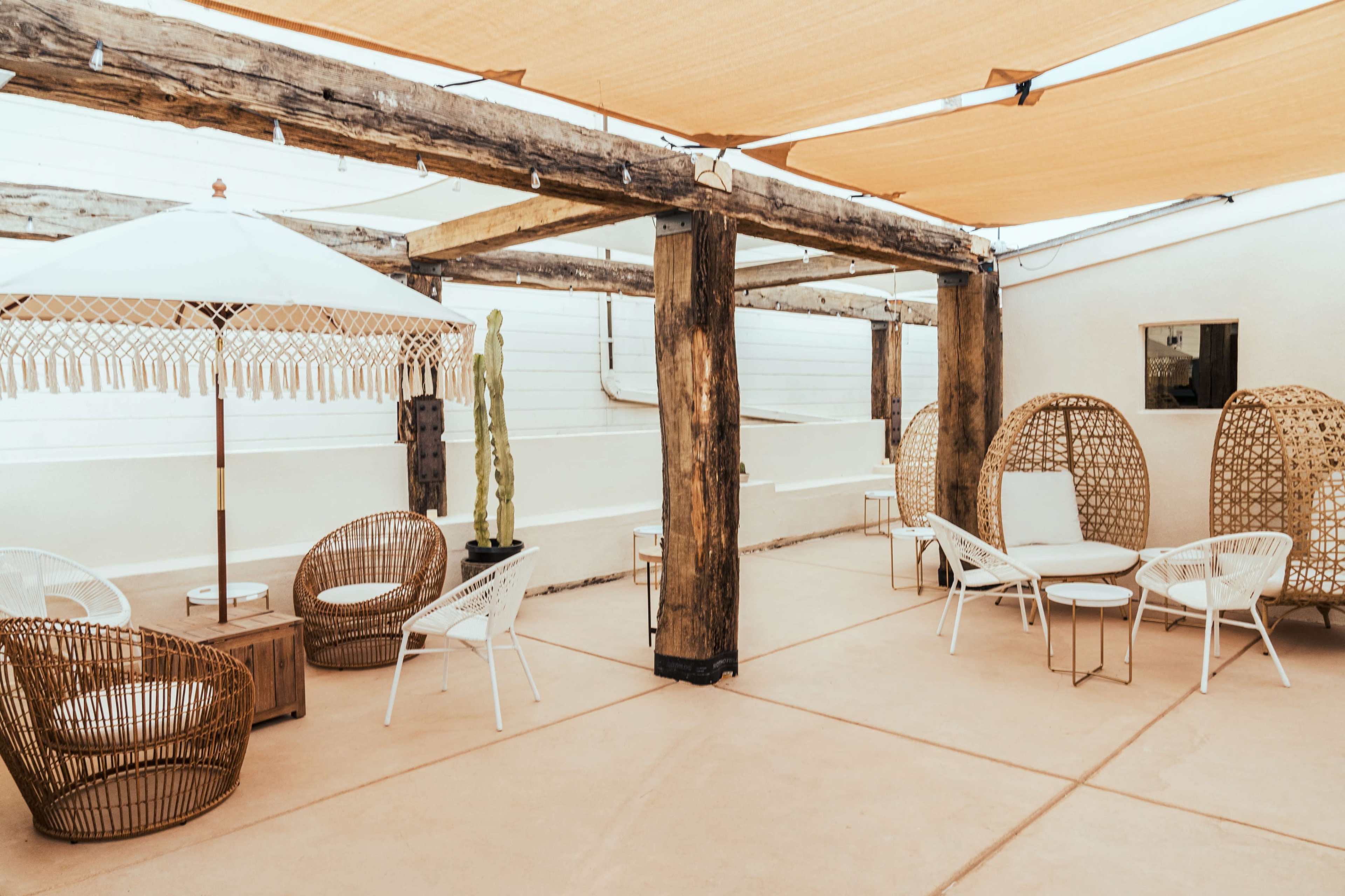 A patio area with wooden pillars, woven seating arrangements, a white umbrella, and a cactus in a minimalistic outdoor setting.