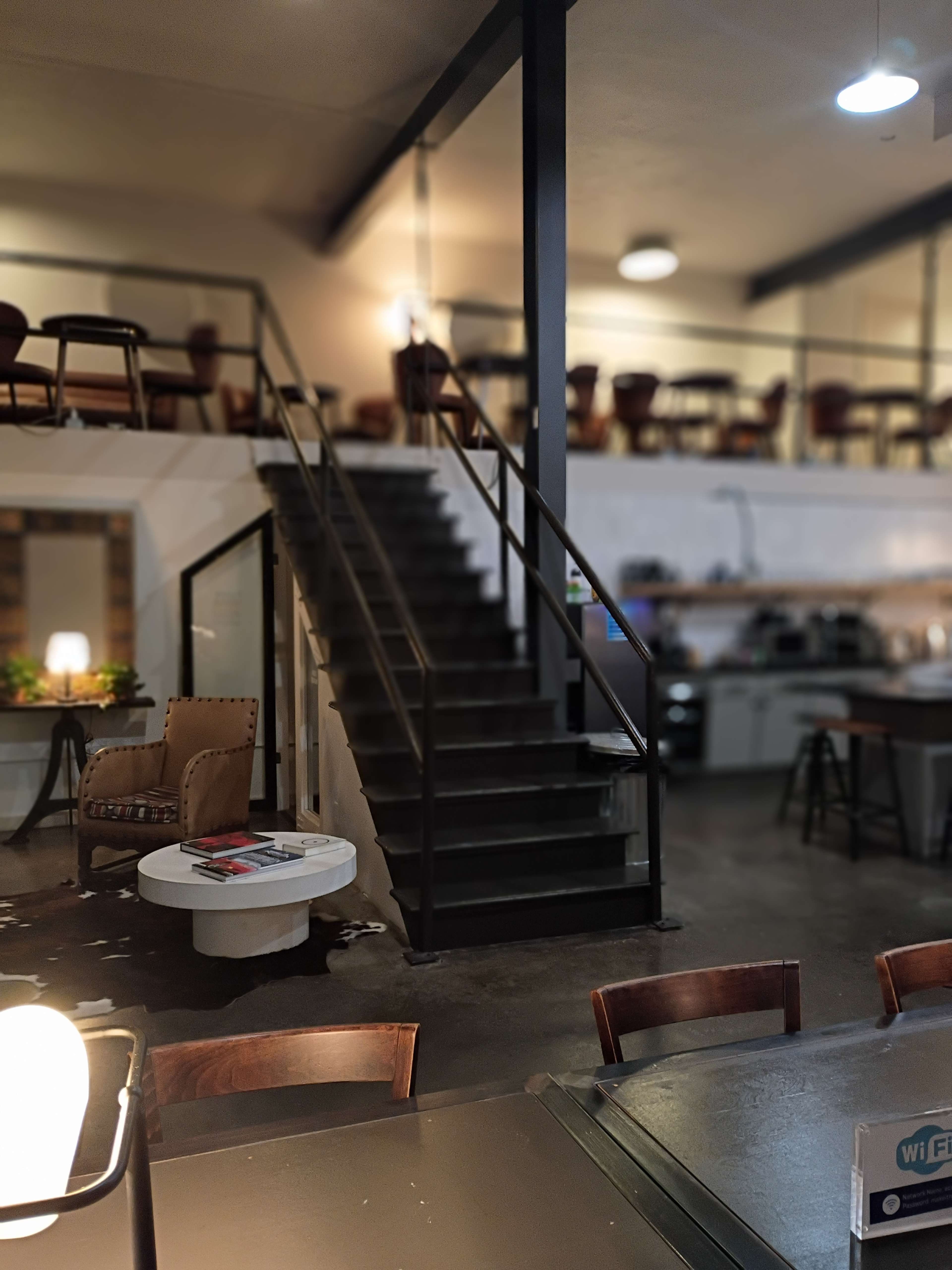 The image shows a modern café interior with a staircase leading to an upper level, surrounded by seating areas and a counter.