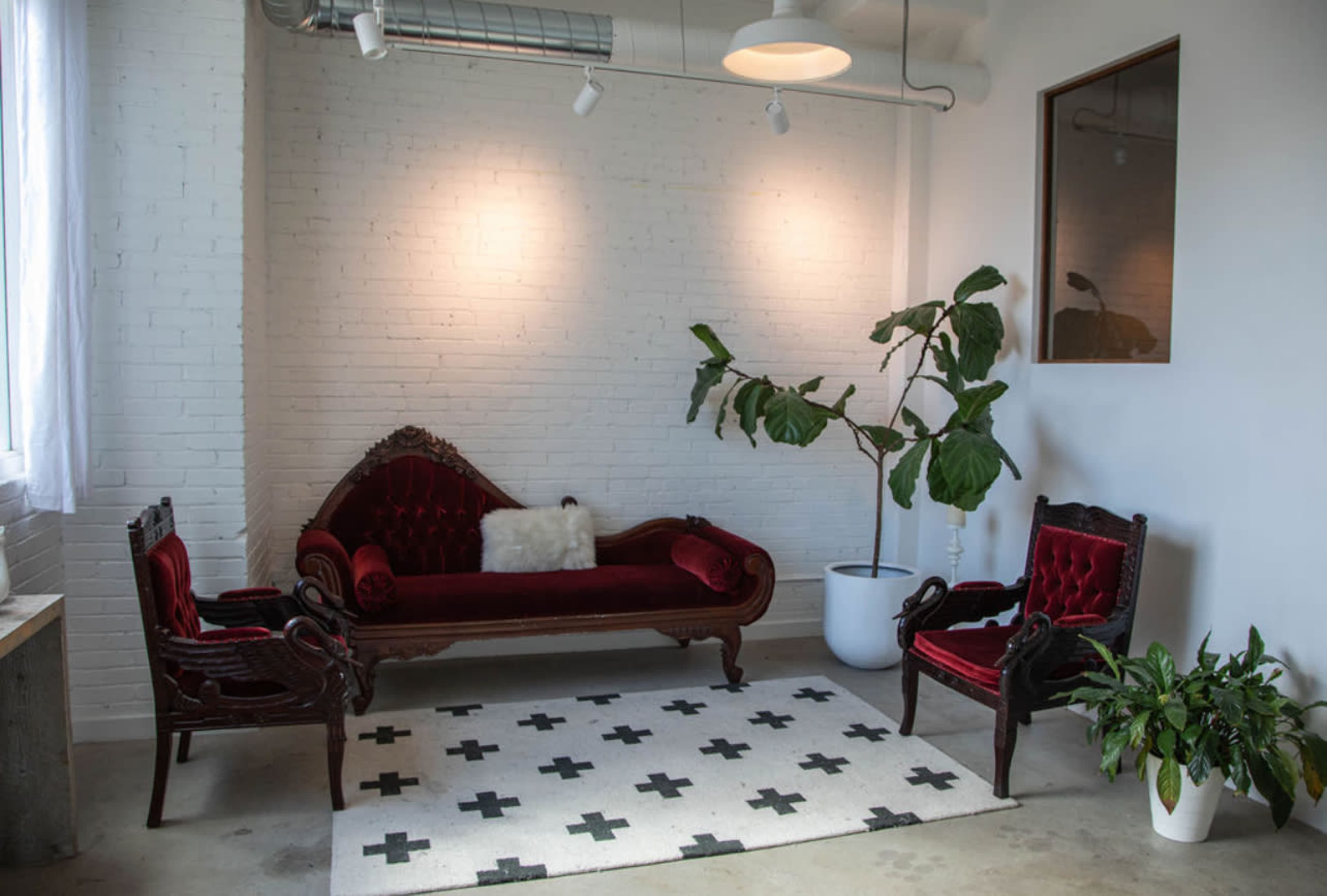 The image shows a seating area with a red velvet sofa, two matching armchairs, a potted plant, and geometric-patterned rug against a white-brick wall.