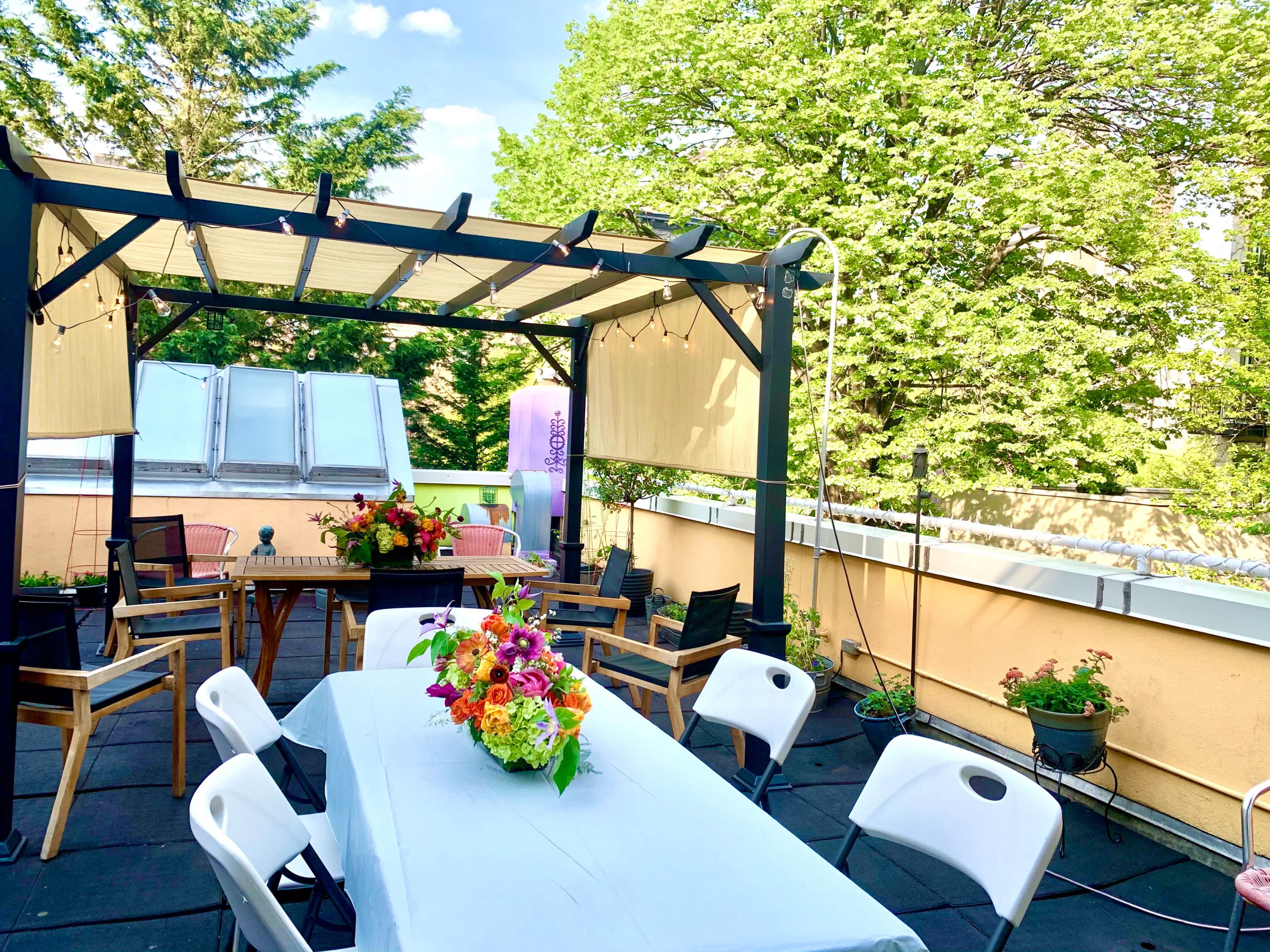 The image shows a rooftop terrace with a dining table set with flowers, surrounded by chairs and potted plants, beneath a canopy.