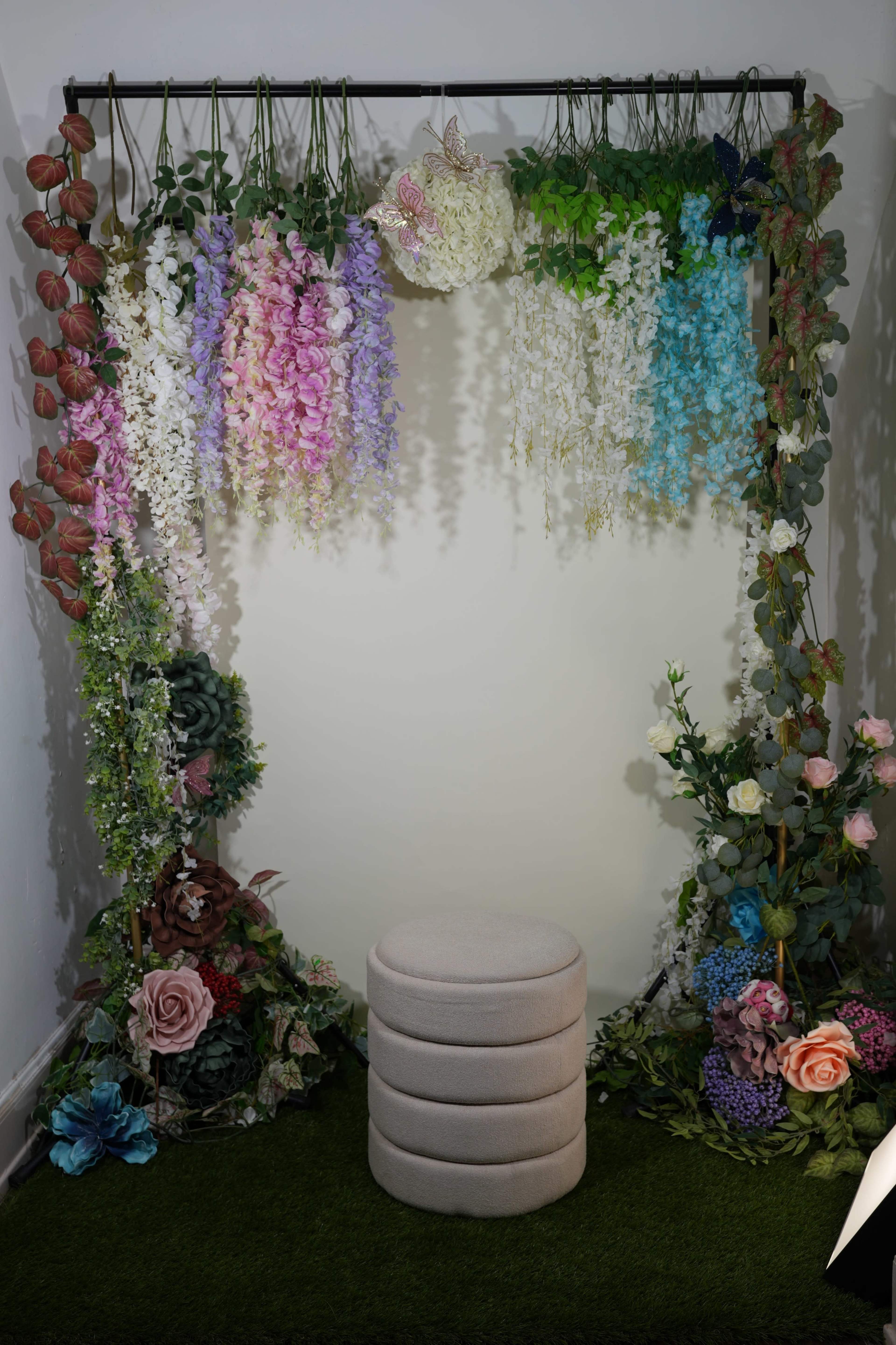 The image shows a decorative backdrop made of various artificial flowers in shades of pink, purple, blue, and white, with a circular stool in front of it.