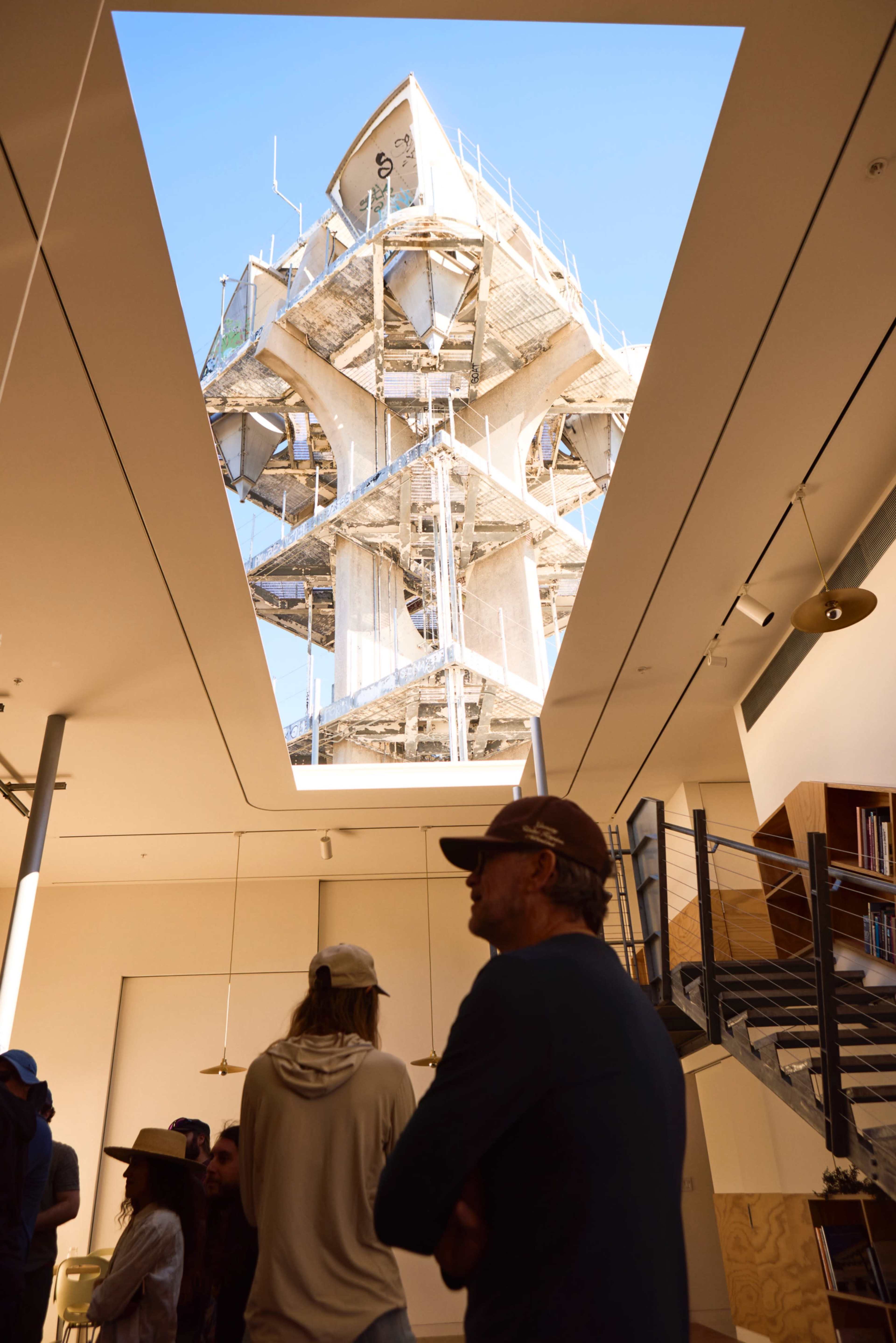 A group of people stands indoors, looking up through a rectangular opening in the ceiling at a construction site featuring a large scaffolded structure.