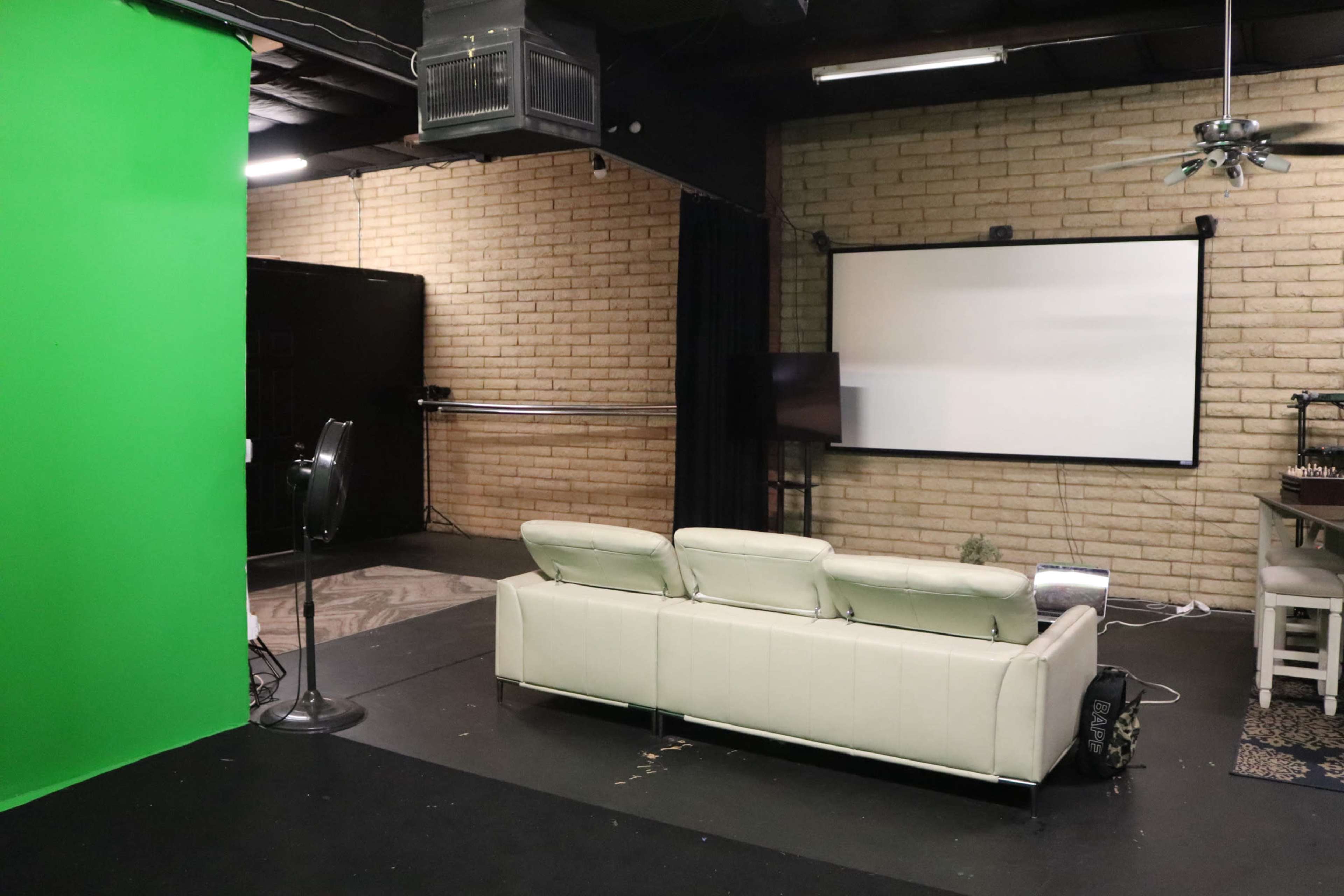 The image shows a studio space featuring a white couch, a green screen, and a large projection screen on a brick wall.