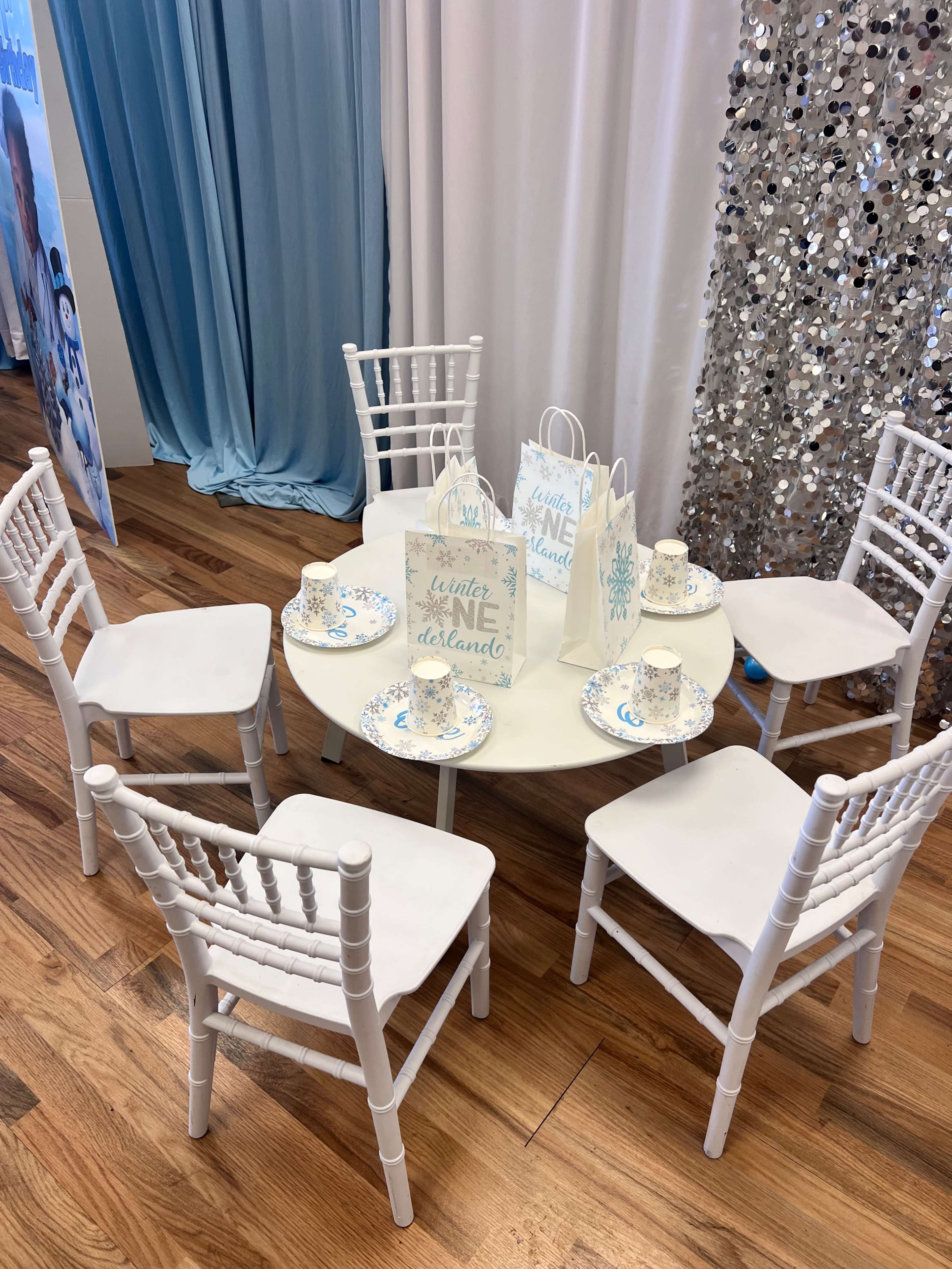 A circular table is set with six white chairs, a decorative bag, and plates with a blue floral pattern in a room with light blue and silver decor.