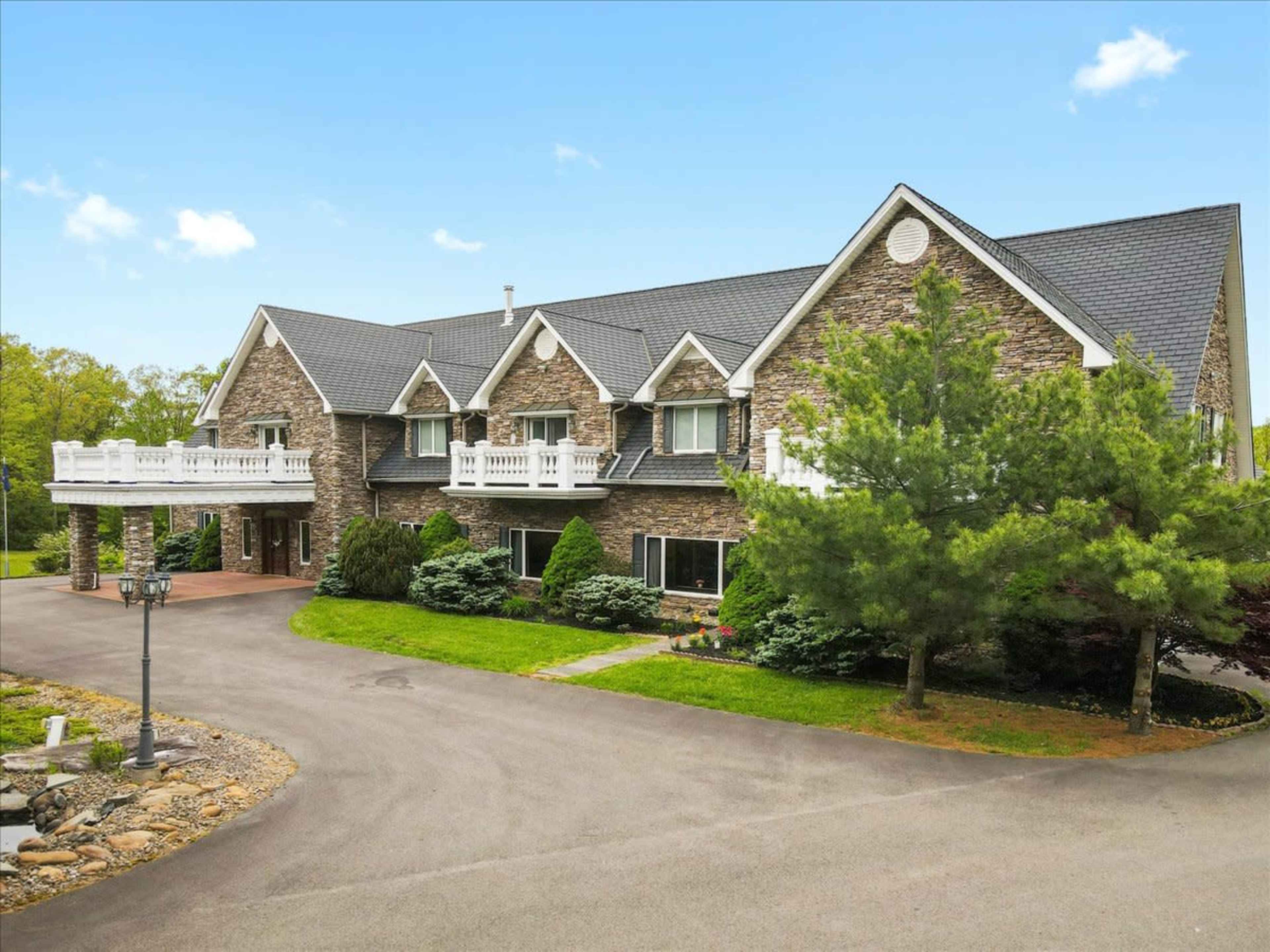 A large stone house with multiple balconies is set along a paved driveway surrounded by landscaped greenery.