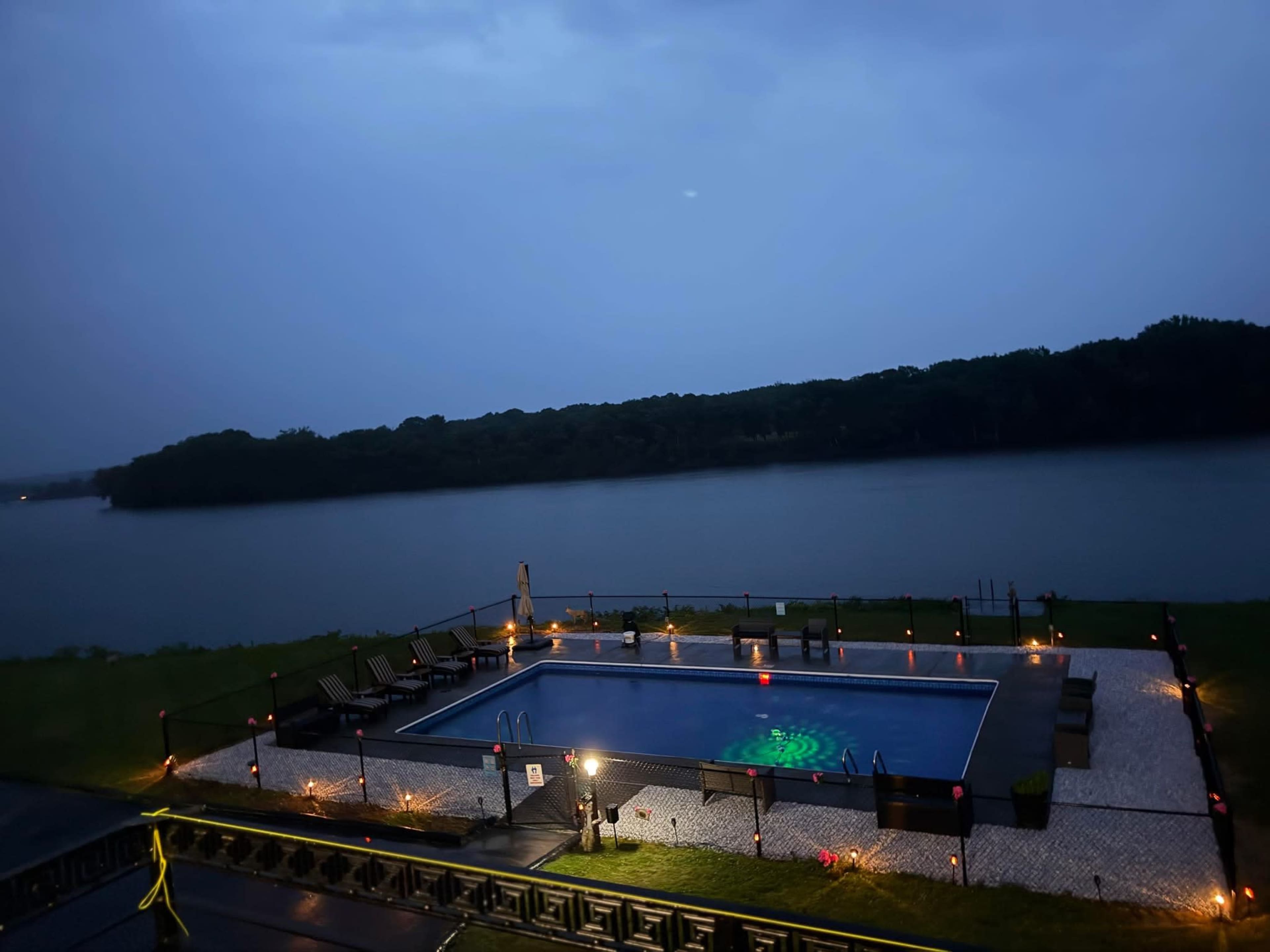 A swimming pool is situated beside a calm lake, illuminated by soft lighting, under a cloudy evening sky.