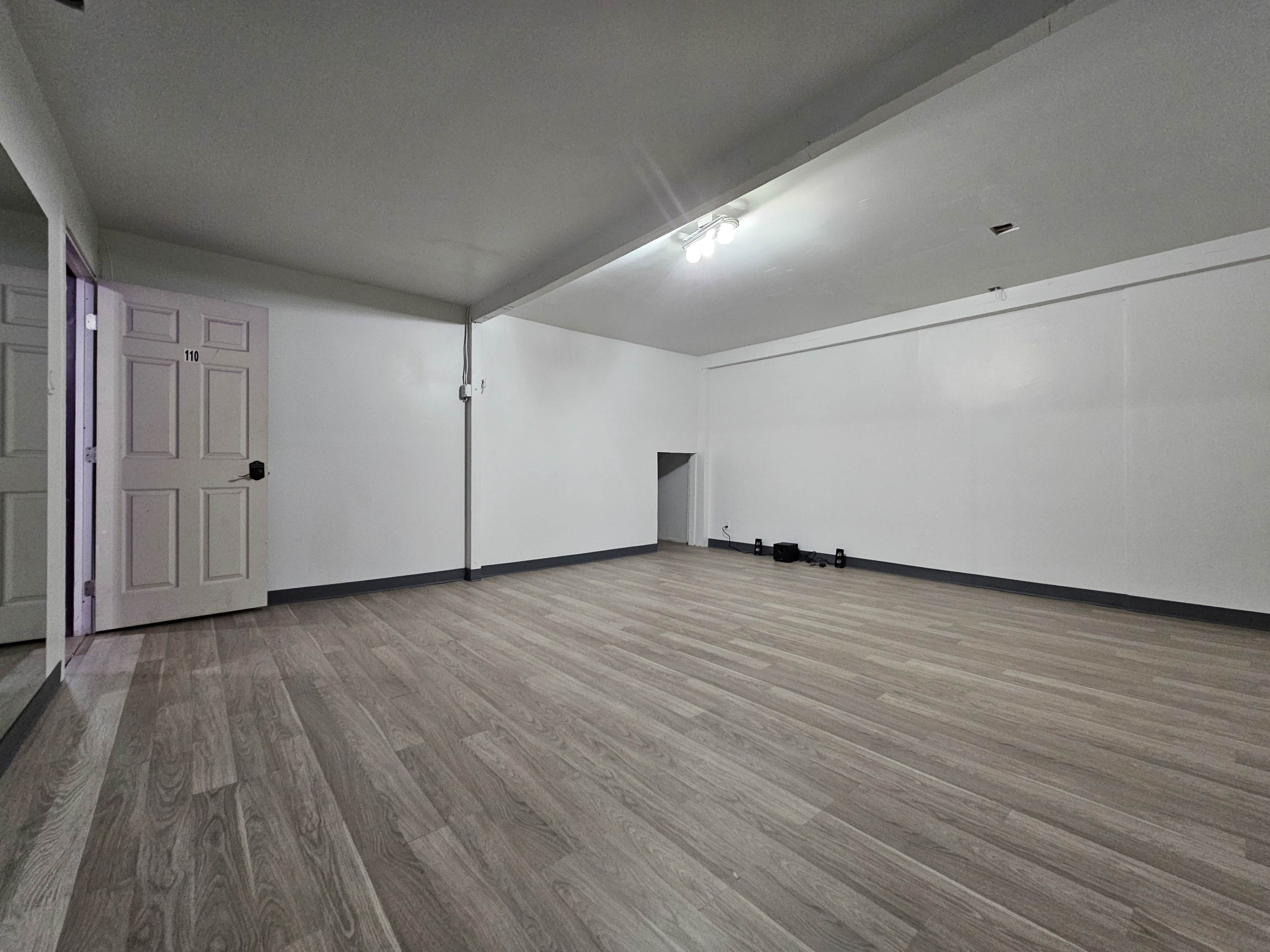 The image shows a spacious, empty room with wooden flooring, a plain white wall, and a door on one side.