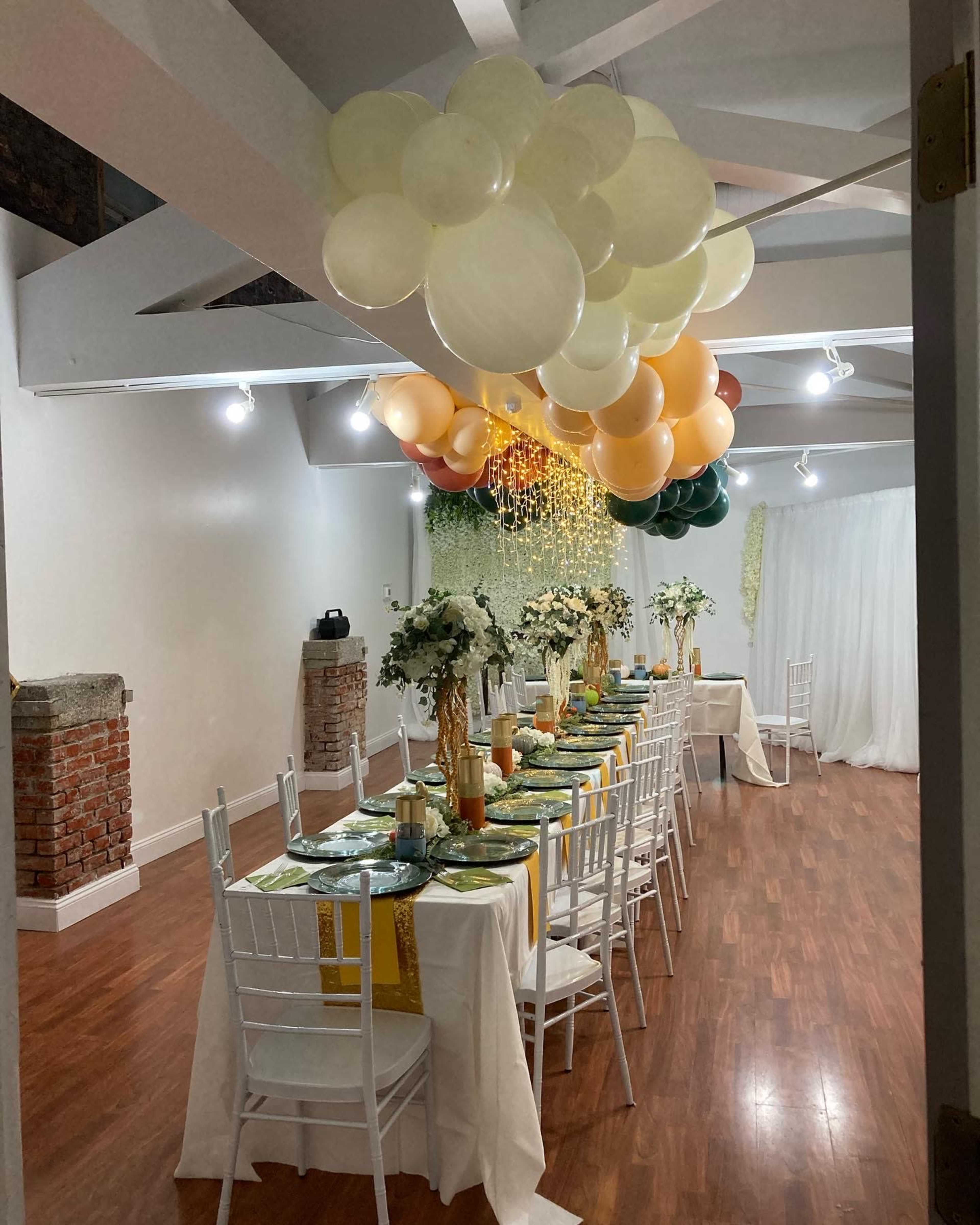 A long dining table is set with green and gold decorations, surrounded by white chairs, beneath a ceiling adorned with a cluster of balloons.