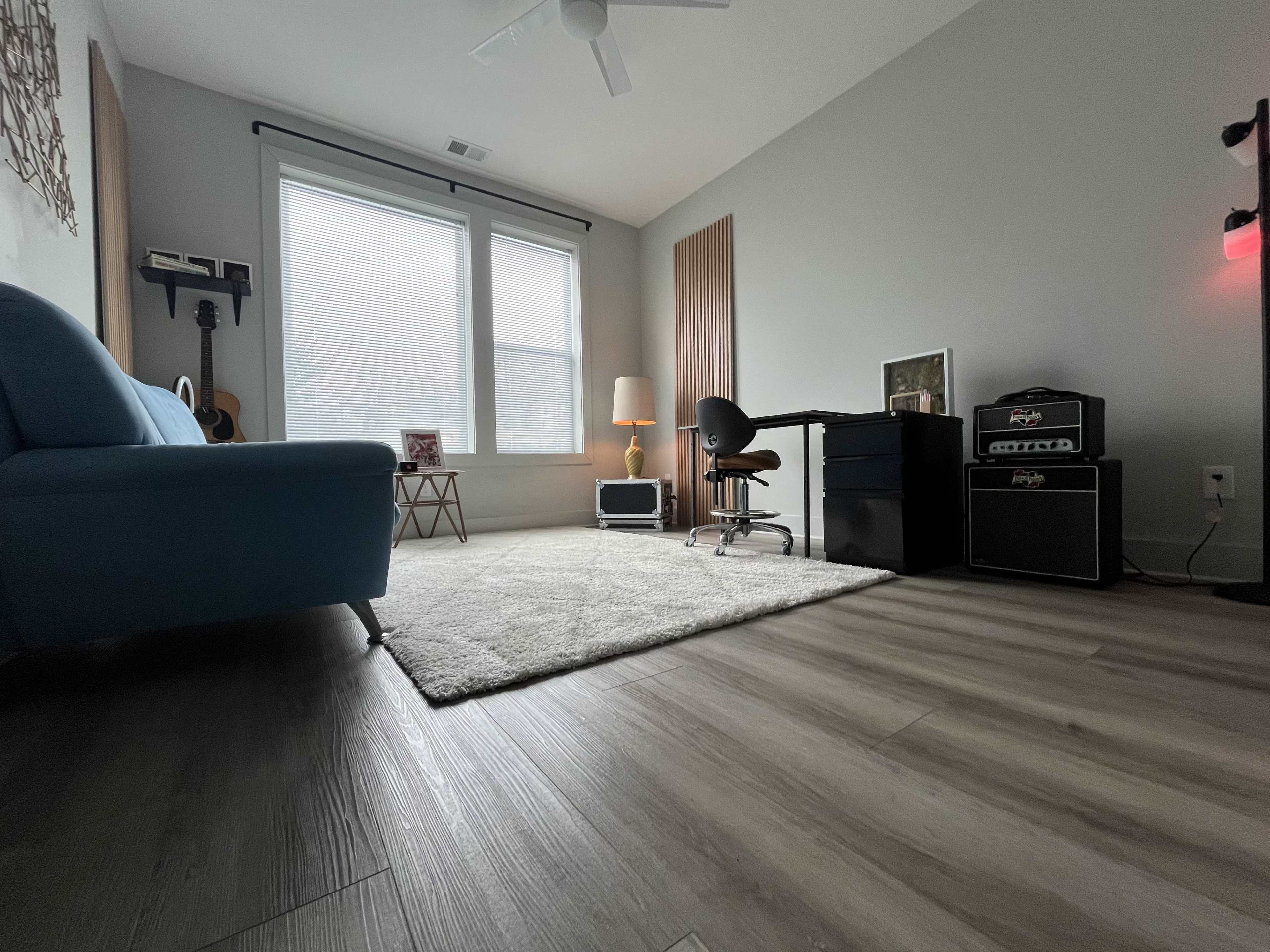 The image shows a spacious room featuring a blue couch, a small side table, a desk with a chair, a tall lamp, and an audio amplifier in one corner.