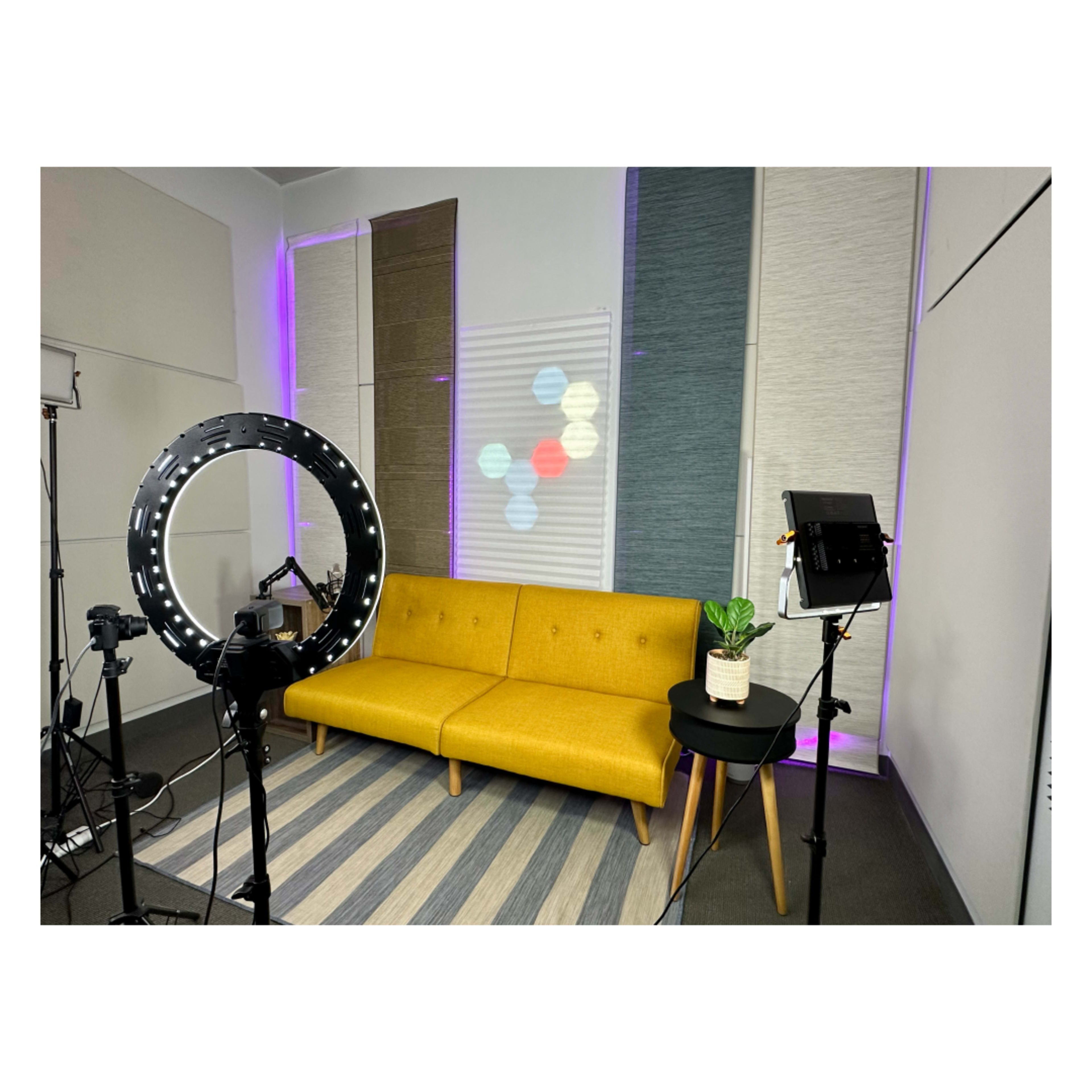 A modern studio setup featuring a yellow sofa, a small black table with a plant, a ring light, and soft lighting creating a colorful backdrop.