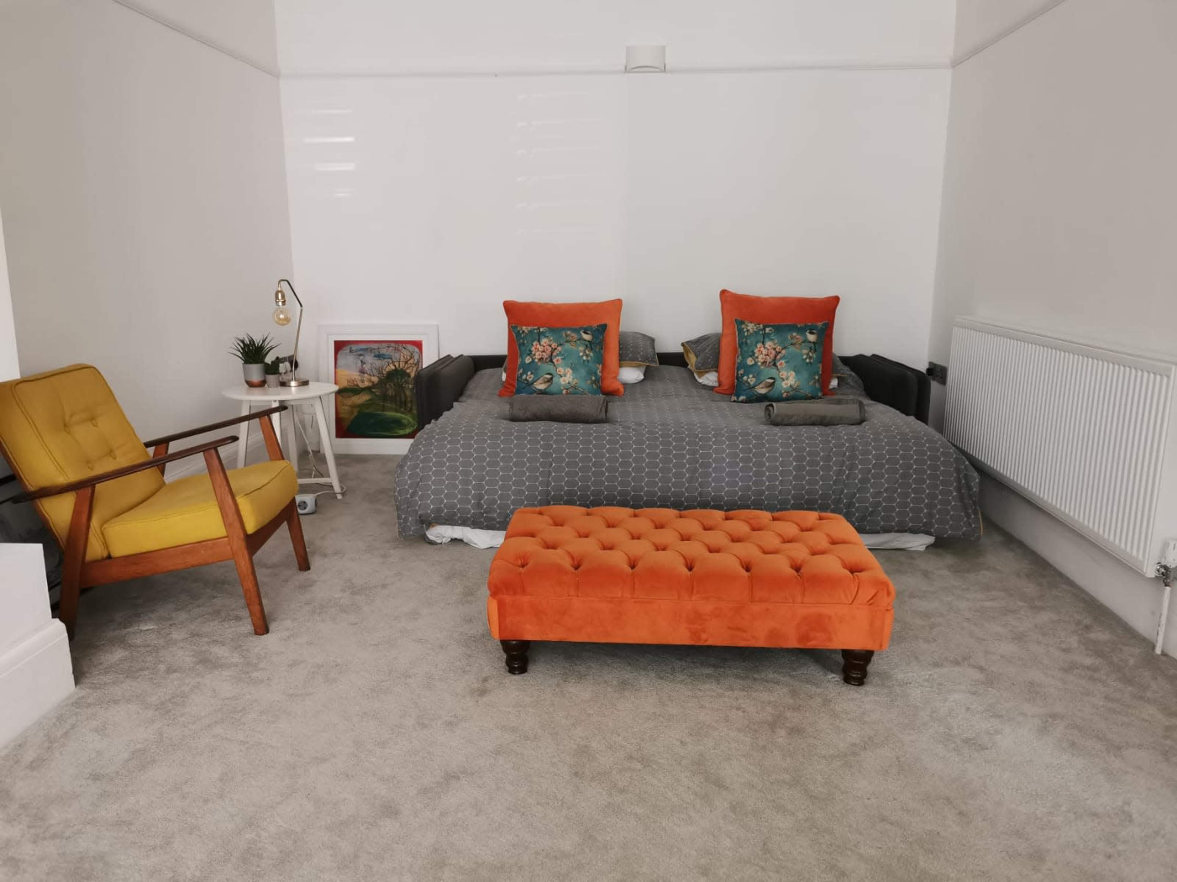 A neatly arranged bedroom features a gray bed with orange and blue accent pillows, an orange upholstered bench, and a yellow armchair beside a radiator.