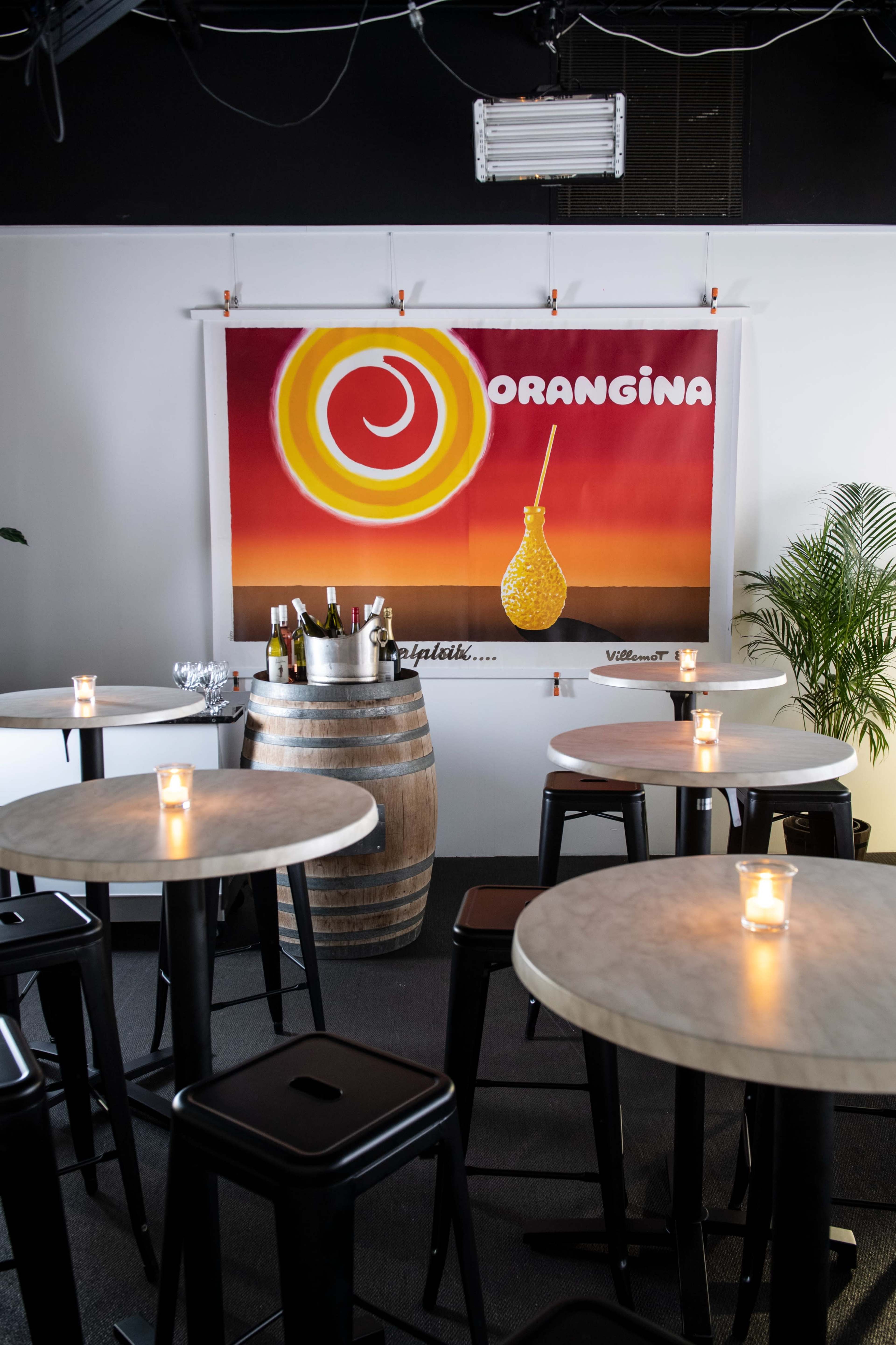 A brightly colored Orangina advertisement hanging on the wall of a casual dining area featuring round tables and a wooden barrel.
