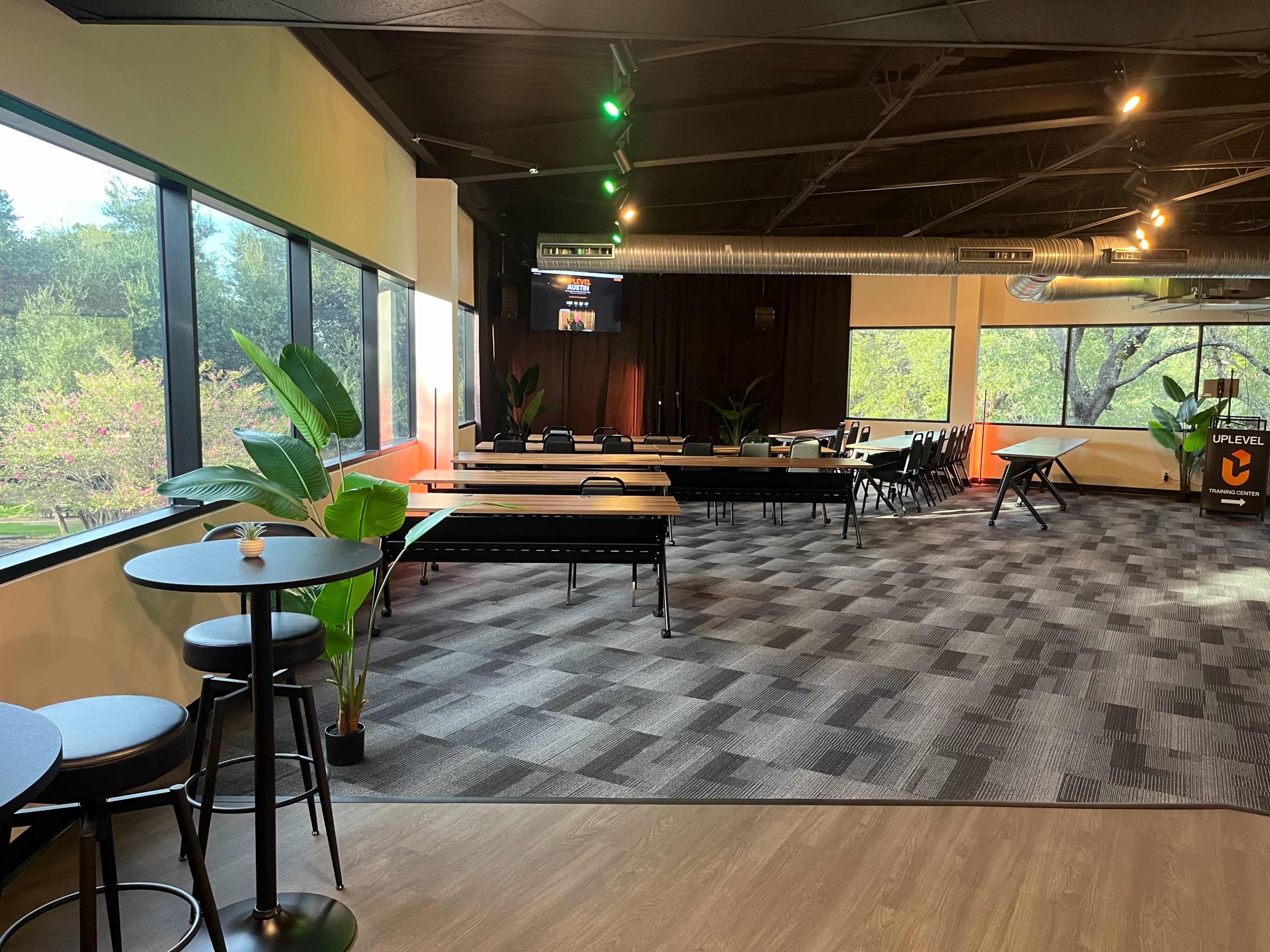 The image shows a spacious indoor area with large windows, featuring tables, seating, and plants, likely intended for social events or gatherings.
