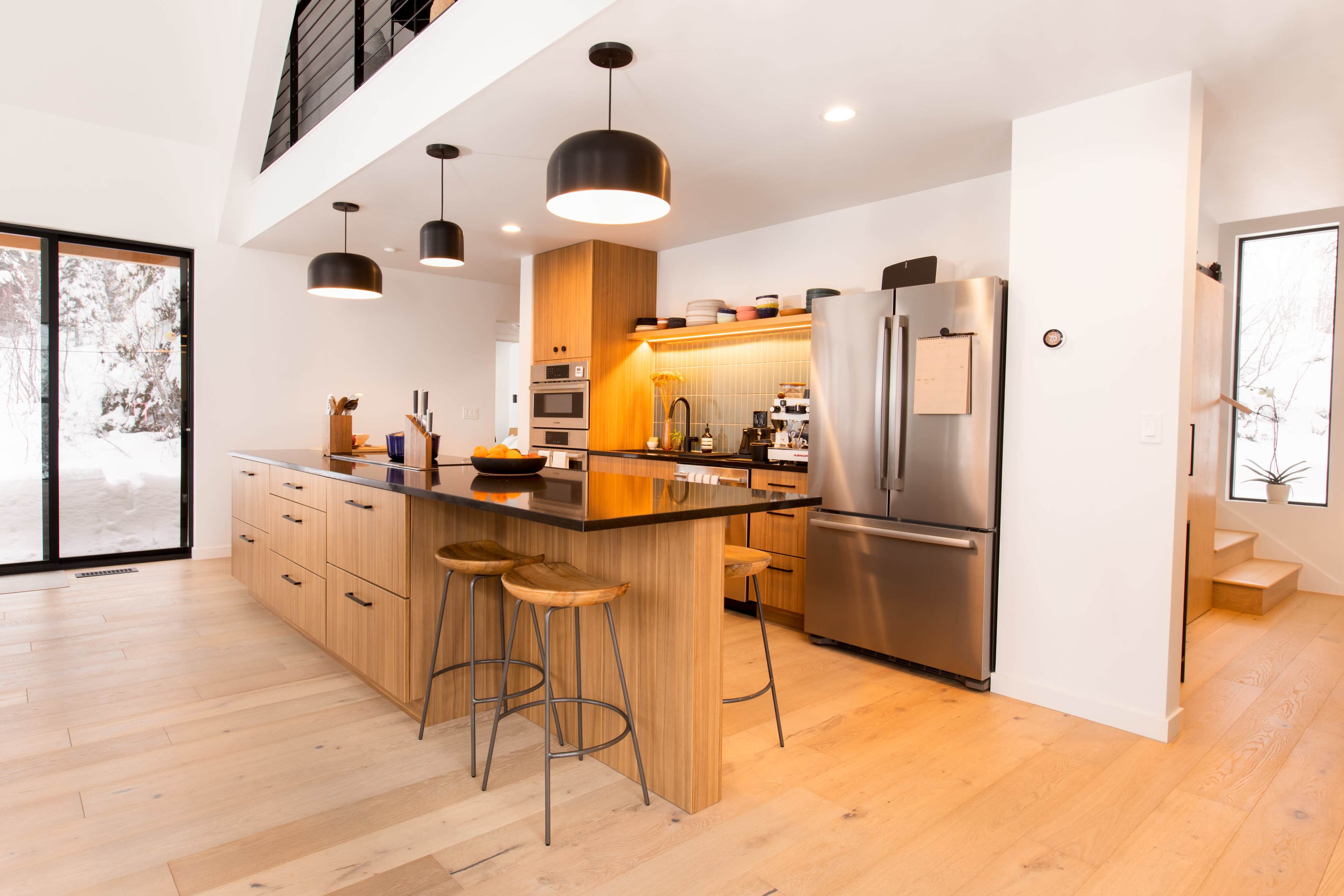 The image shows a modern kitchen with a large island, wooden cabinetry, stainless steel appliances, and ample natural light.
