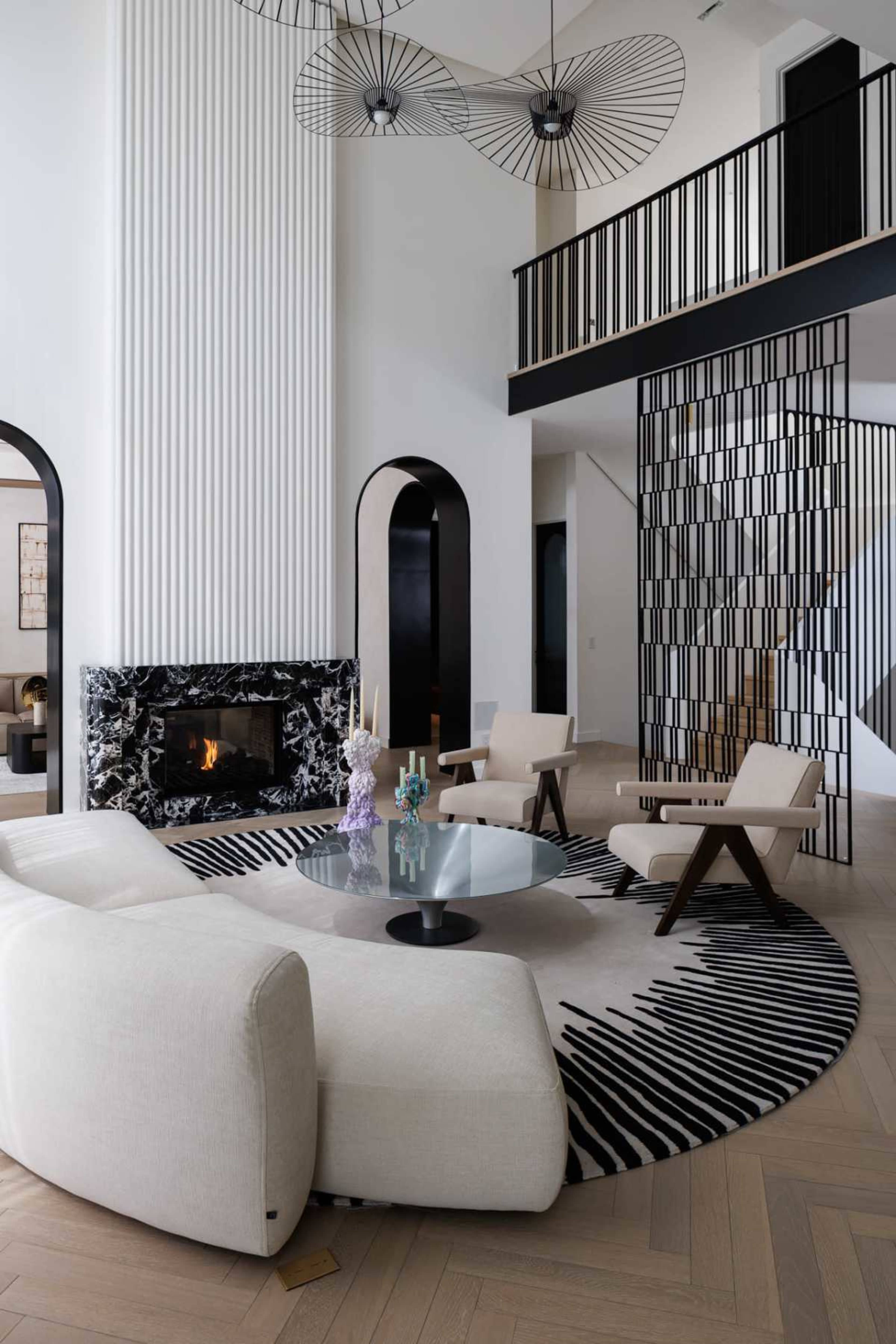 The image features a modern living room with a curved white sofa, a round black and white patterned rug, a glass coffee table, and a black marble fireplace, all complemented by tall ceilings and striking architectural features.