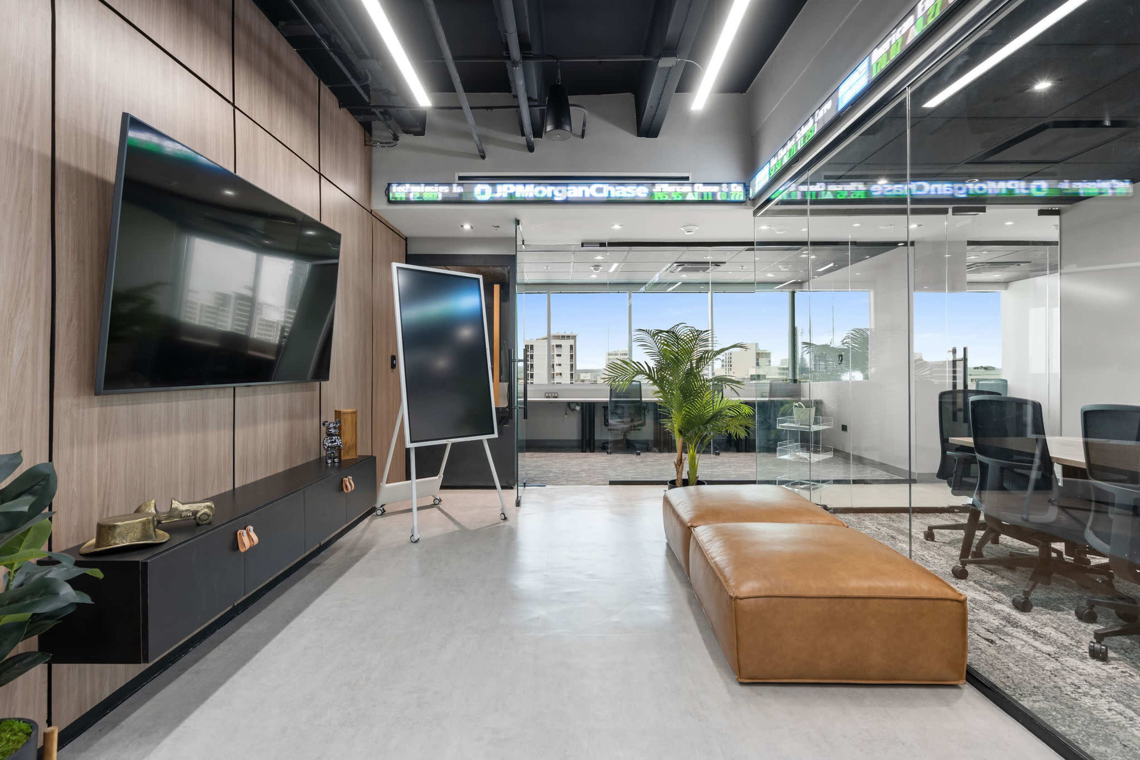 The image shows a modern office space with a television mounted on a wooden wall, a large screen displaying stock information, and a brown bench opposite a glass-walled conference room.