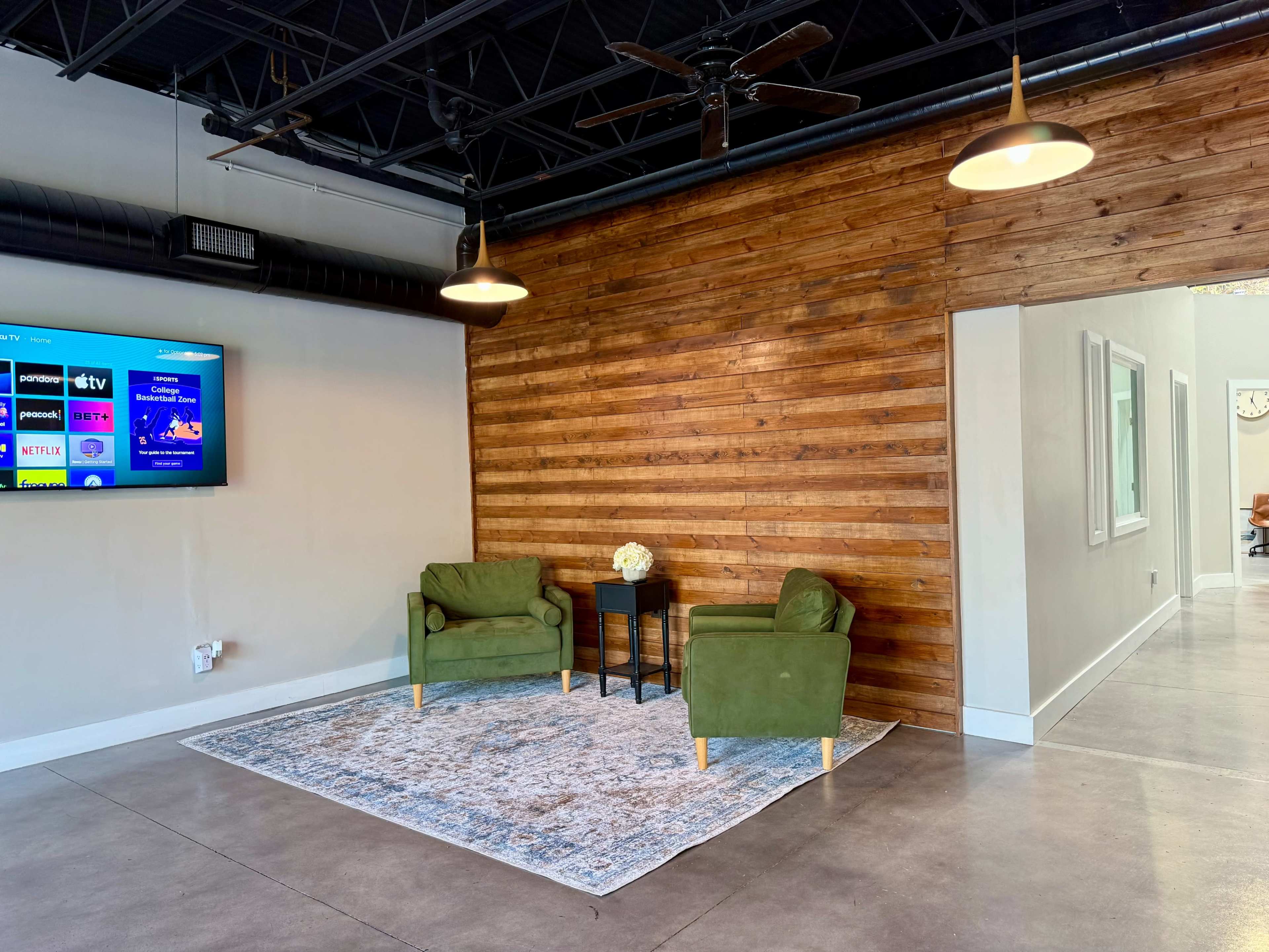 The image shows a modern interior space with two green chairs, a small table, and a wooden feature wall, illuminated by pendant lights.