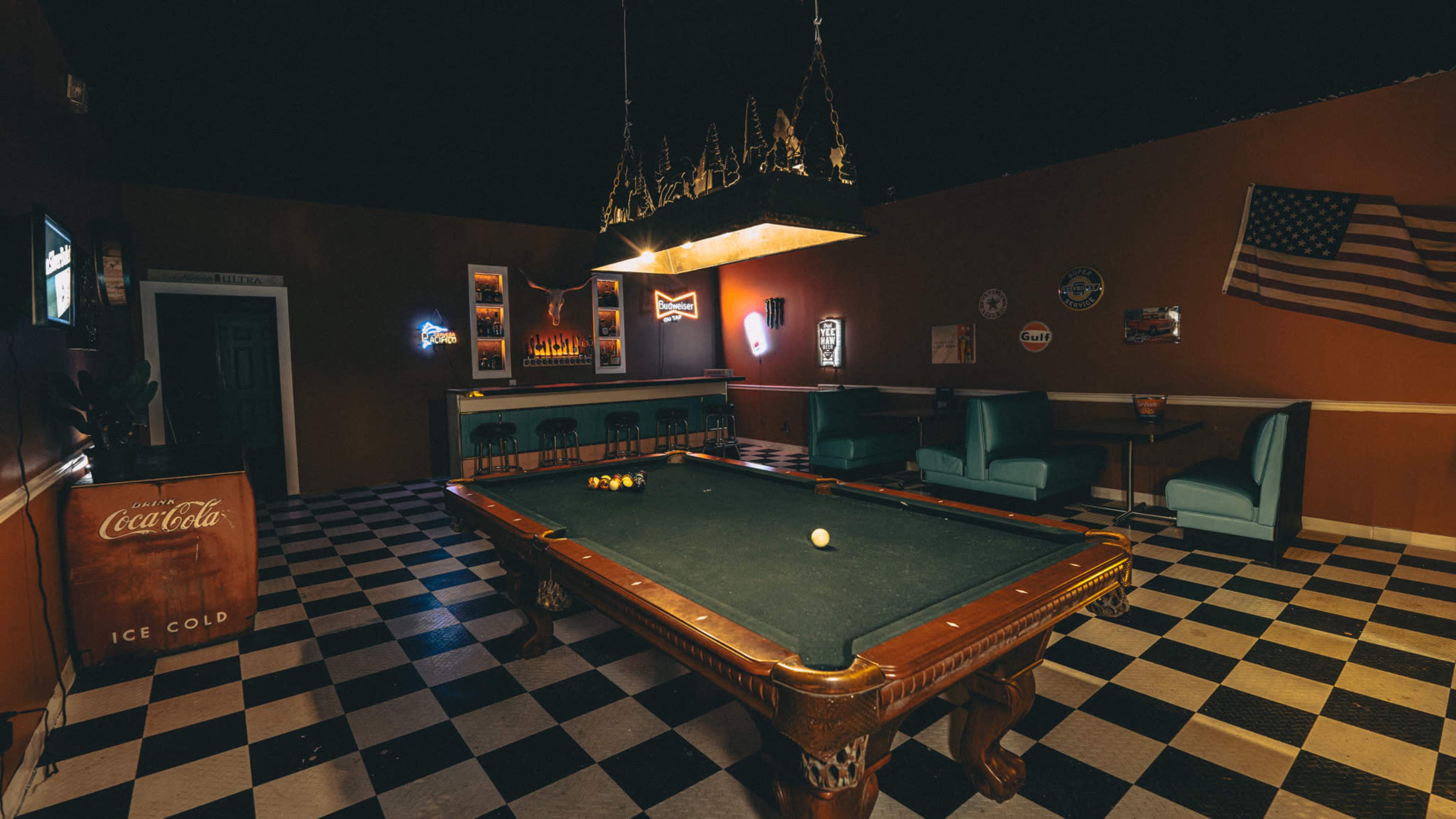 The image shows a dimly lit room featuring a billiards table at the center, surrounded by teal seating and adorned with vintage signs and an American flag on the wall.